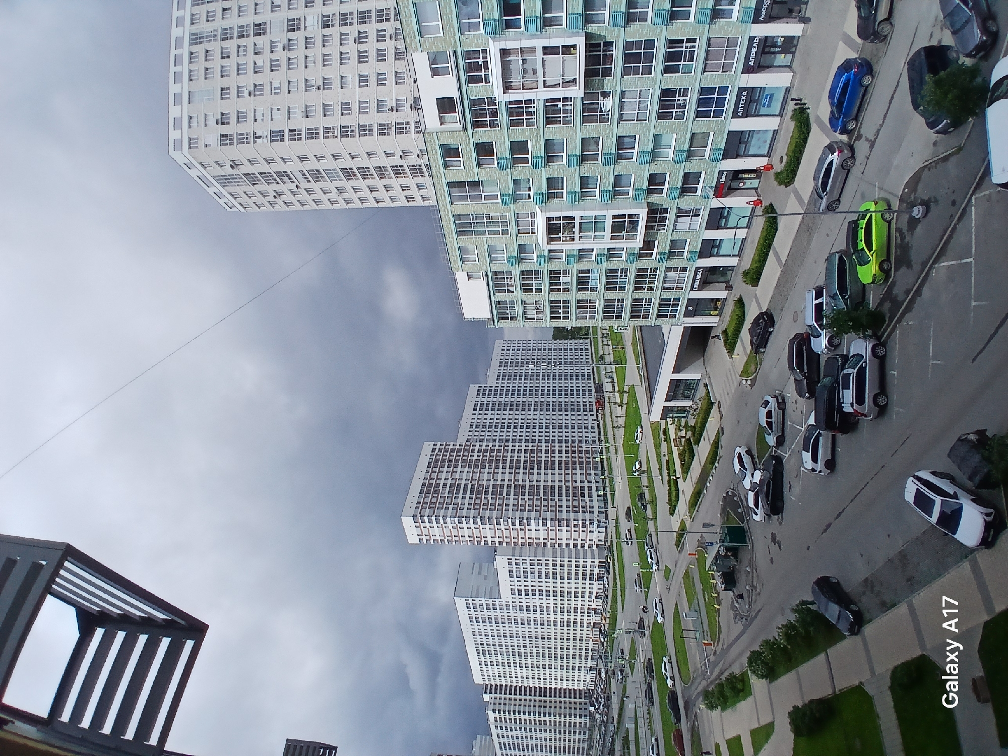 The image is a wide-angle, high-angle shot of a modern urban residential area in Yekaterinburg, Russia. The scene is dominated by several multi-story apartment buildings, some of which are very tall and white, while others are lower and clad in light teal and white. A network of roads and parking lots is visible between the buildings, with numerous cars parked and some in motion. The sky is overcast with dark, heavy clouds, suggesting inclement weather. In the foreground, a portion of a balcony structure from a building is visible in the upper left corner. The ground level shows pedestrian walkways, green spaces, and the entrances to shops and businesses located on the ground floors of the teal and white building, with visible signage in Russian. Cars include sedans, SUVs, and hatchbacks in various colors. No individuals are clearly identifiable or engaged in notable activity. The overall impression is of a densely populated, contemporary residential district under a dark, cloudy sky.