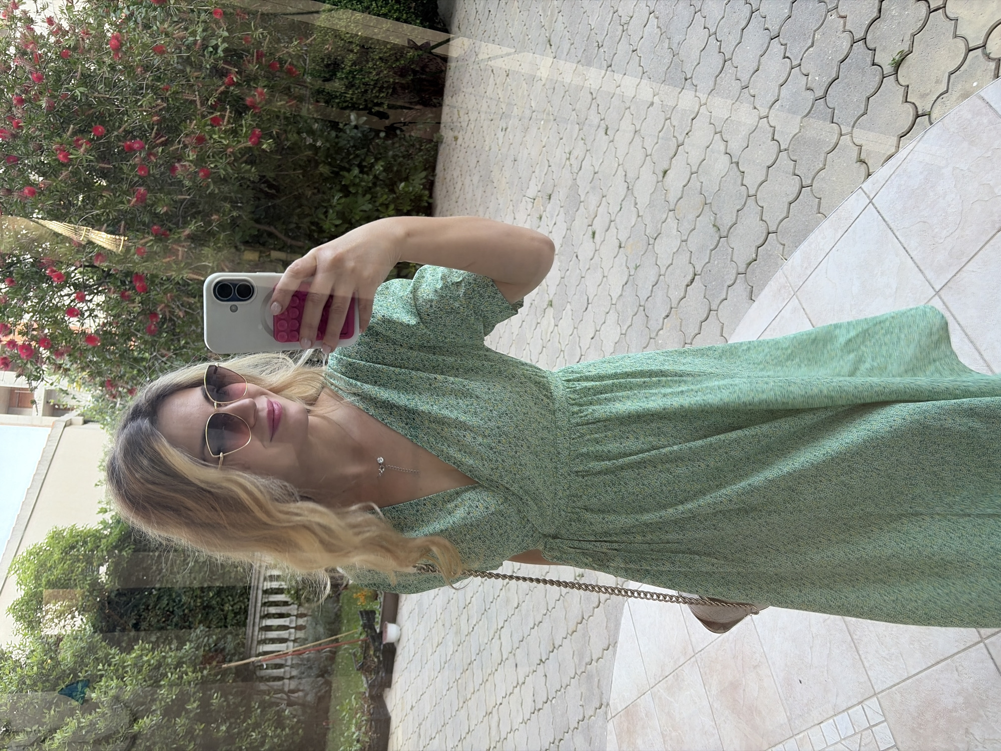 A blonde woman is taking a selfie in front of a window. She is wearing a light green dress and sunglasses. The window reflects a garden with trees and red flowers. The background shows a paved area with stone patterns and greenery, possibly in Budva, Montenegro, given the typical Mediterranean architecture and flora. The woman is holding a smartphone in her right hand.