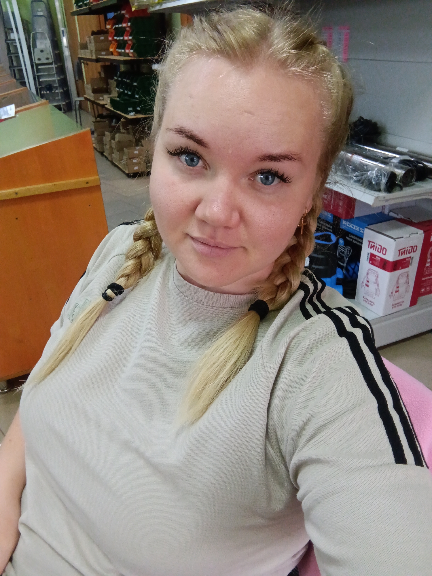 A medium shot depicts a woman with fair skin and blonde hair styled in braids. She is wearing a light-colored, long-sleeved shirt with black stripes on the sleeves. The woman has blue eyes and appears to be indoors, likely in a commercial space. The background includes shelves stocked with various items, indicating a store setting. The image was taken in the city of Фоки, Russia.