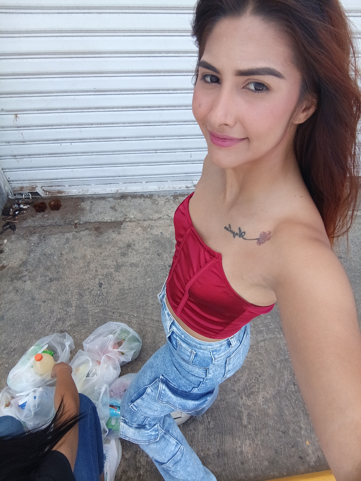 An outdoor, high-angle shot captures a woman looking directly at the camera with a gentle smile. She appears to be in her 20s or 30s, with shoulder-length reddish-brown hair and fair skin. She is wearing a strapless, form-fitting red top and light blue, acid-wash jeans. A tattoo is visible on her left collarbone, featuring the word "Always" in a script font, followed by a small, stylized flower or leaf design.
The background is a light grey or white corrugated metal roll-down door or shutter, suggesting a commercial building or storage unit. The ground is rough, dirty concrete.
In the lower left portion of the image, the arm, leg, and dark hair of a second person are partially visible, indicating they are seated on the ground. This person is also wearing blue jeans and a black top. Scattered around this second individual are several clear plastic bags, appearing to contain groceries or recently purchased items. Visible contents include a bottle with an orange cap, partially labeled "LEMONADE", and another bottle with a blue label, partially reading "BONICE".
The scene is brightly lit, suggesting it is daytime. The overall impression is a casual moment, possibly after shopping, in an urban or semi-urban environment consistent with Juan Griego, Venezuela.