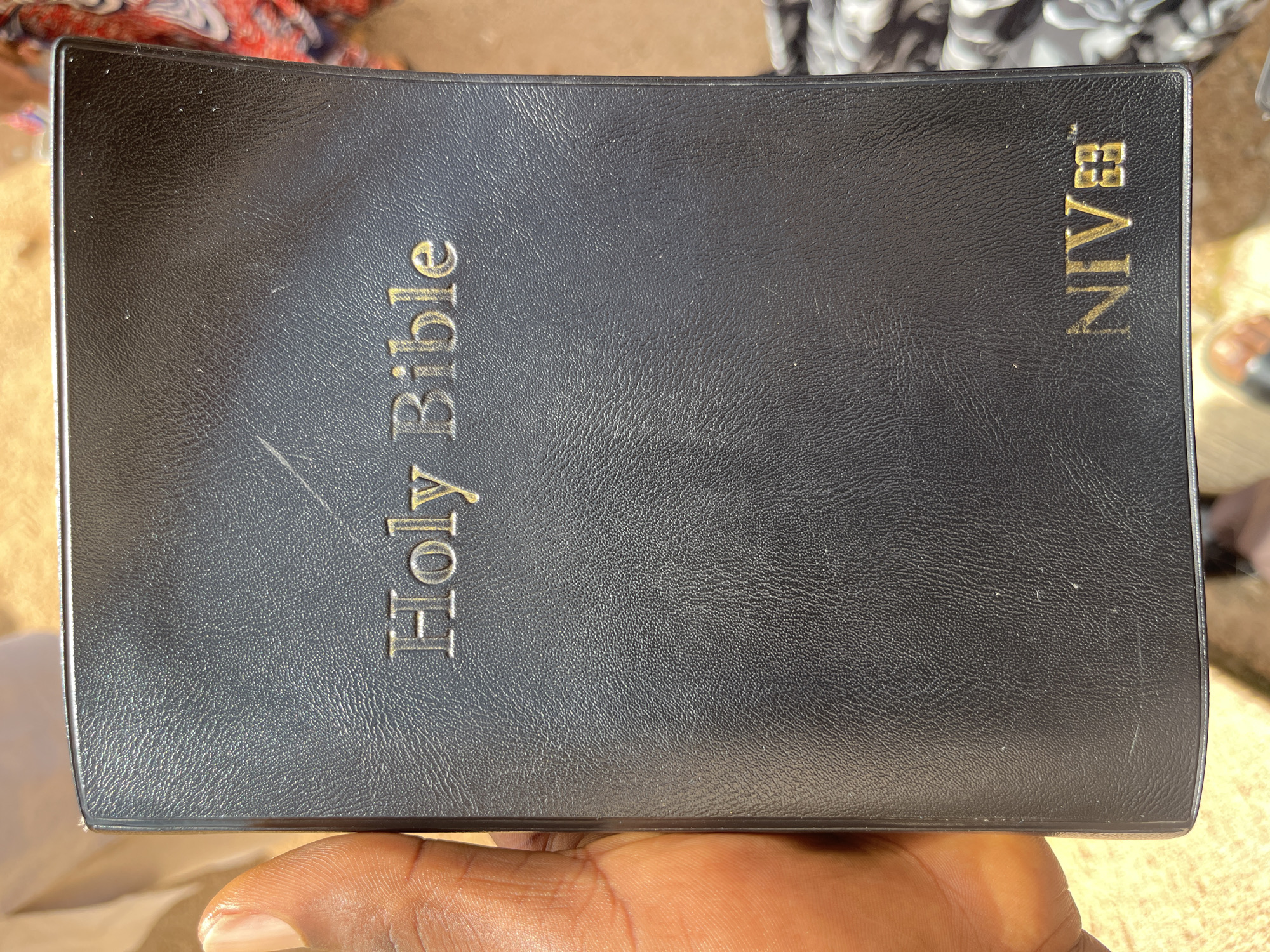 A close-up, overhead shot shows a person's thumb and index finger holding the corner of a black, leather-bound Holy Bible. The Bible is embossed with gold lettering on the cover that reads "Holy Bible" in an elegant font, and the "NIV" logo is visible in the bottom right corner. The Bible is positioned vertically, with the spine facing to the left. The background is blurred, showing a light brown surface and hints of patterned fabric. The scene is set outdoors, likely in Bukuru, Nigeria, under natural daylight. The overall impression is one of religious devotion and personal connection with the sacred text.