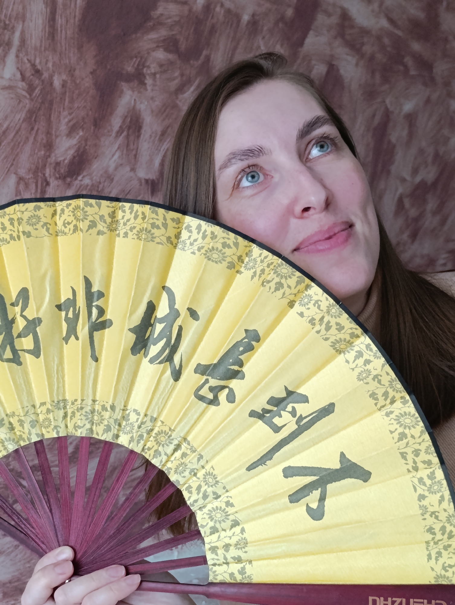 A woman with fair skin and blue eyes is holding a yellow and maroon Chinese fan. The fan has black Chinese characters written on it. The woman is looking upwards and to the right. The background is a mottled brown. The woman is wearing a neutral-colored turtleneck sweater. The location of Perm, Russia is not discernible from the image.