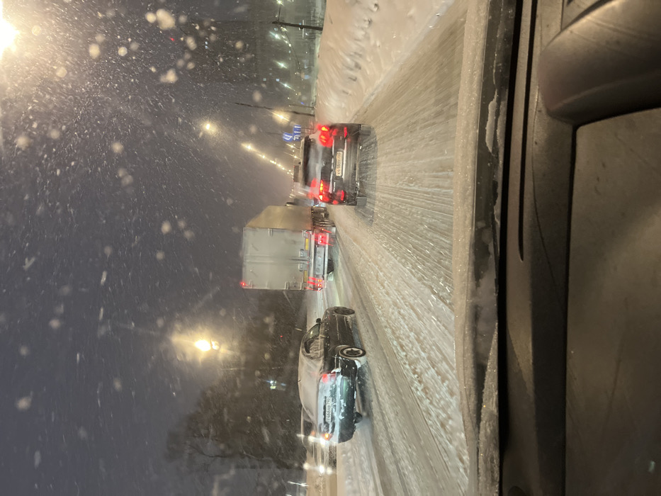 A forensic examination of the image reveals a nighttime street scene in Solnechnogorsk, Russia, during heavy snowfall. The perspective is from the interior of a vehicle, looking forward through a windshield, with the dashboard visible in the foreground, showing a light dusting of snow or frost. The multi-lane road ahead is heavily covered in slush and packed snow, displaying numerous tire tracks. Traffic is present, with vehicles proceeding or idling.
In the left visible lane, a dark sedan, possibly a Chevrolet Lacetti, with accumulated snow on its rear, displays Russian license plate T466HB 750. Immediately to its right, a large white semi-truck with a box trailer travels in the same direction, its taillights illuminated. To the right of the semi-truck and slightly closer to the viewer, a dark SUV, possibly a Kia Sportage, shows illuminated taillights and bears Russian license plate Y418BY 750.
The environment is illuminated by numerous bright streetlights lining the road and by the headlights and taillights of vehicles, which highlight the dense, falling snowflakes. A significant bank of snow forms along the right side of the road. No individuals are directly visible.