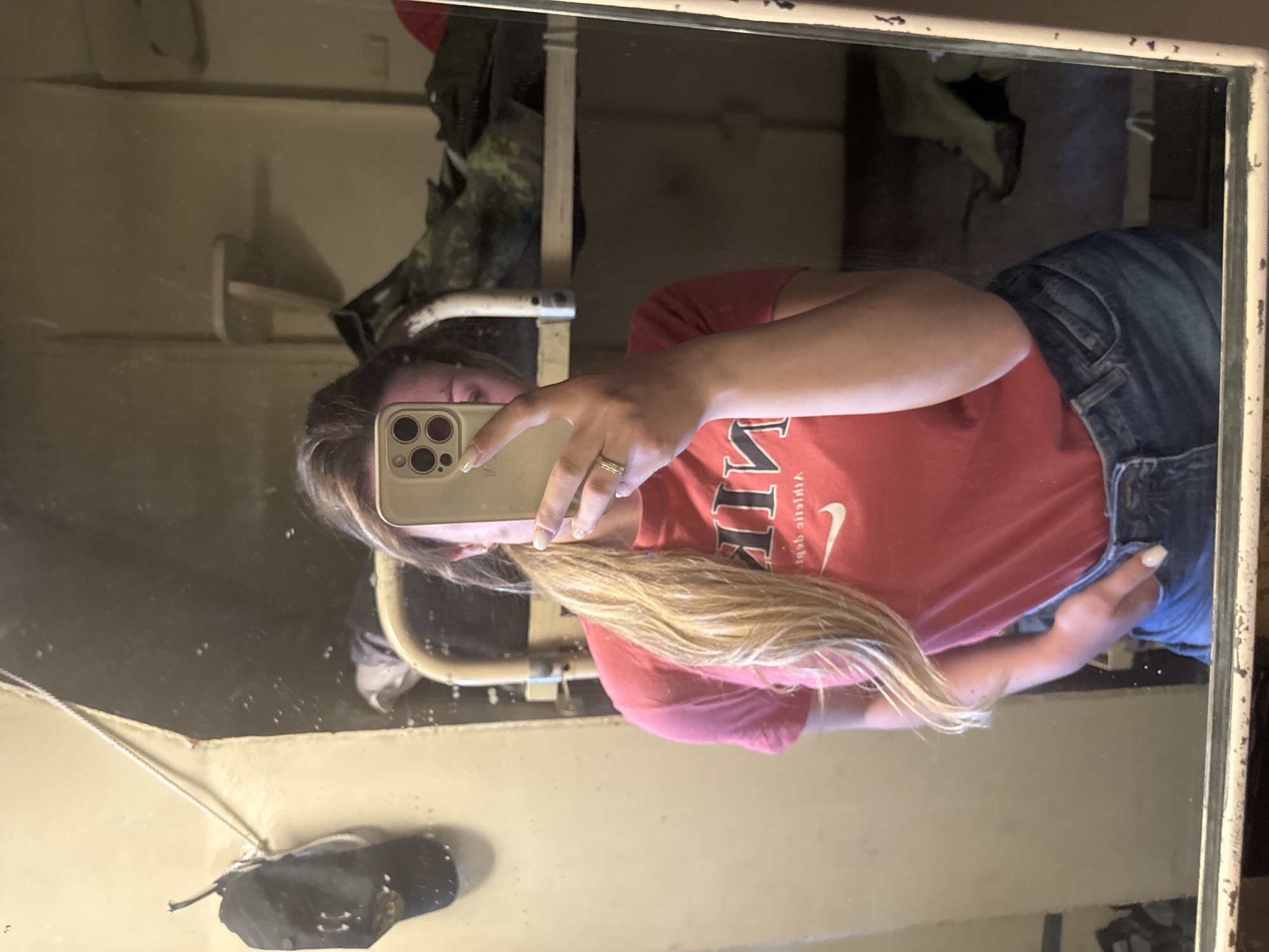 A woman is taking a mirror selfie. She is wearing a red t-shirt and blue jeans. Her hair is blonde. She is holding a smartphone in her left hand. The background includes a shelf with items on it, and the mirror frame is worn. There is a black face mask on the lower left corner of the photo. It is not possible to determine the location in Varna, Bulgaria from the provided image.