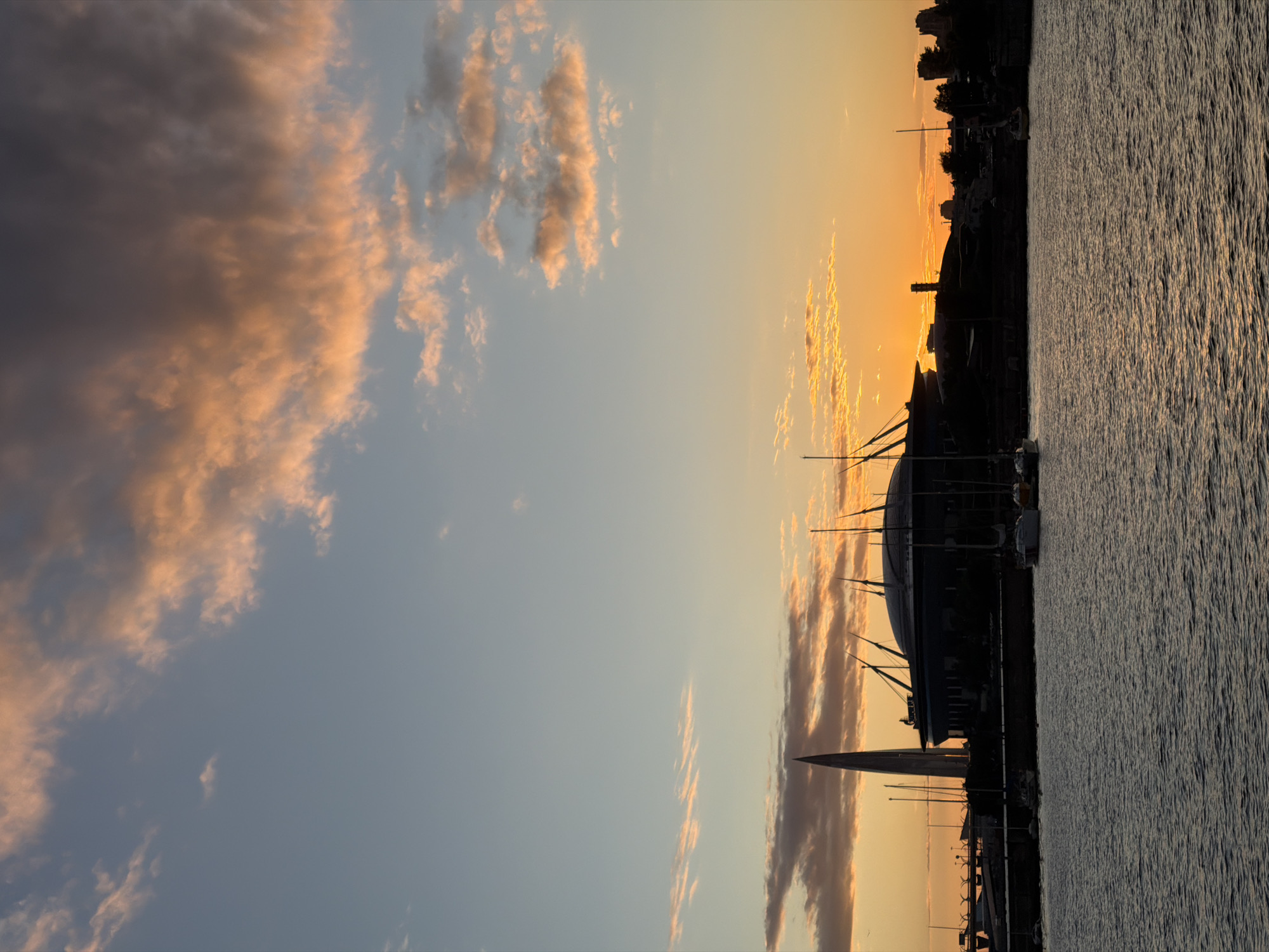 This image captures a sunset view of the Gazprom Arena and the Lakhta Center tower in Saint Petersburg, Russia, as seen from the Neva River. The sky is a gradient of blue and orange, with scattered clouds tinged pink and gold by the setting sun. Silhouetted against the vibrant sky are the distinct architectural features of the arena, a large domed structure, and the tall, needle-like Lakhta Center. Several sailboats are docked or moored in the foreground, their masts also silhouetted. The water of the Neva River is dark and rippled, reflecting the colors of the sky. The overall atmosphere is serene and picturesque, showcasing a prominent urban landscape during a beautiful sunset.