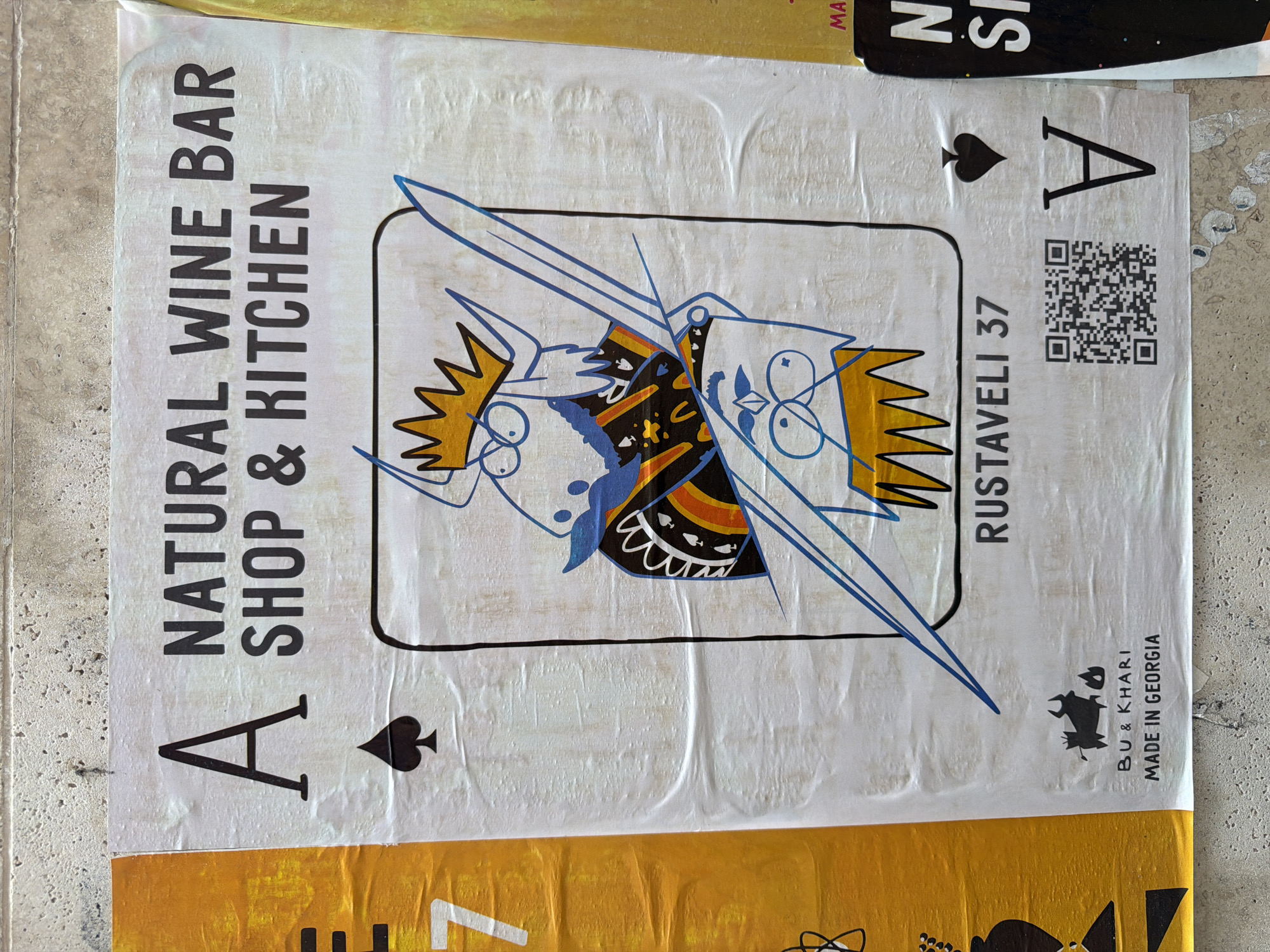 The image shows a poster for a "NATURAL WINE BAR SHOP & KITCHEN" located at Rustaveli 37 in Tbilisi, Georgia. The poster is designed to resemble a playing card, specifically the Ace of Spades. The central graphic features a cartoon-like illustration of two figures, possibly a king and queen, dressed in ornate clothing and holding a sword. One figure wears a golden crown and glasses, while the other has blue eyes and a diamond shape on its forehead. Below the illustration, the text "RUSTAVELI 37" is clearly visible. At the bottom left of the poster, there is a logo of a bull with horns and the text "BU & KHARI MADE IN GEORGIA". To the right of the logo, a QR code is present. The poster is weathered and slightly torn, attached to a textured concrete wall, suggesting it has been displayed outdoors for some time. Another poster, predominantly yellow with the number "7" prominently displayed, is visible on the left, and a portion of a black poster with white text is seen on the right.