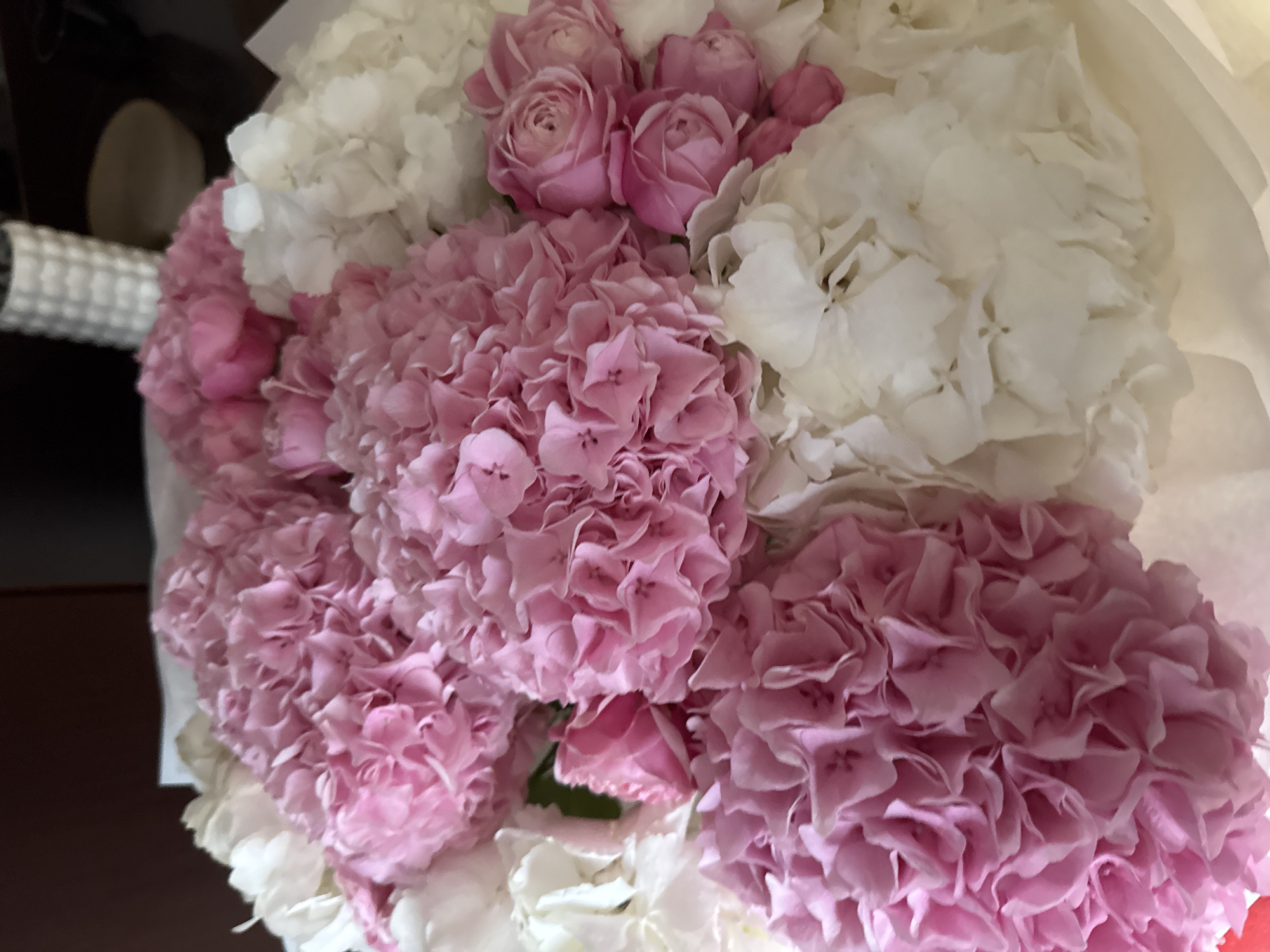 The image displays a close-up, top-down view of a beautiful floral arrangement, likely a bouquet, observed in Warsaw, Poland. The primary subjects are fresh flowers, predominantly in shades of soft pink and pristine white.
Large, full clusters of delicate pink hydrangeas are prominent, characterized by their numerous small, crinkled petals. These pink blooms are interspersed with equally large clusters of white hydrangeas, providing a soft contrast. Nestled among the pink hydrangeas, particularly in the upper-middle section, are several small, tightly budded pink roses, adding a different form and texture to the arrangement. The flowers appear fresh and are densely packed, filling most of the frame.
The background is dark and out of focus, suggesting an indoor setting. On the far left, a blurry, light-colored object with a textured or ribbed surface is partially visible, which may be part of a vase or decorative wrapping. No specific activity or event is depicted; the image is a static portrayal of the flowers. There are no visible people, specific time of day cues, weather details, or discernible text. The arrangement conveys an overall sense of delicate beauty and freshness.