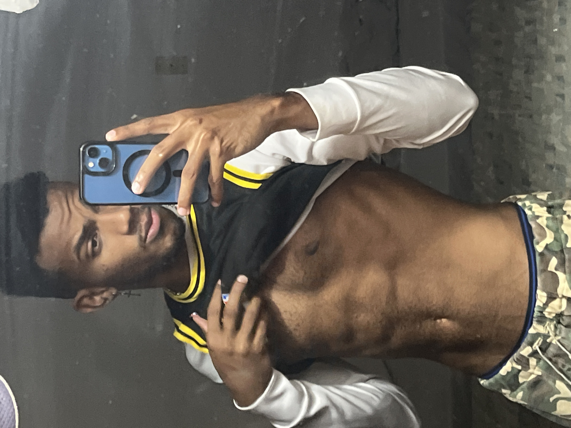 A dark-skinned man is taking a selfie in what appears to be a mirror. He is wearing a black jersey with yellow trim over a white long-sleeved shirt, and camouflage patterned shorts with a blue waistband. The man is holding a blue iPhone in his left hand, partially obscuring his face. The background is a plain, gray wall. There is no visible information to determine a location in Porlamar, Venezuela.