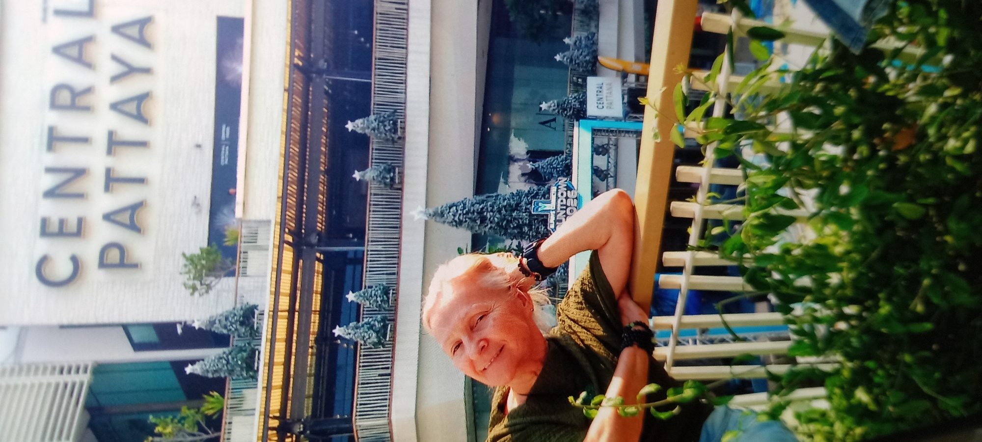 A woman poses in front of the Central Pattaya shopping center in Baan Chaloklum, Thailand. The woman, fair-skinned, wears an olive-green shirt and has wristbands on both wrists. She is leaning on a trellis covered in green foliage. The shopping center is decorated with Christmas trees. In the background, the words "CENTRAL PATTAYA" are visible.