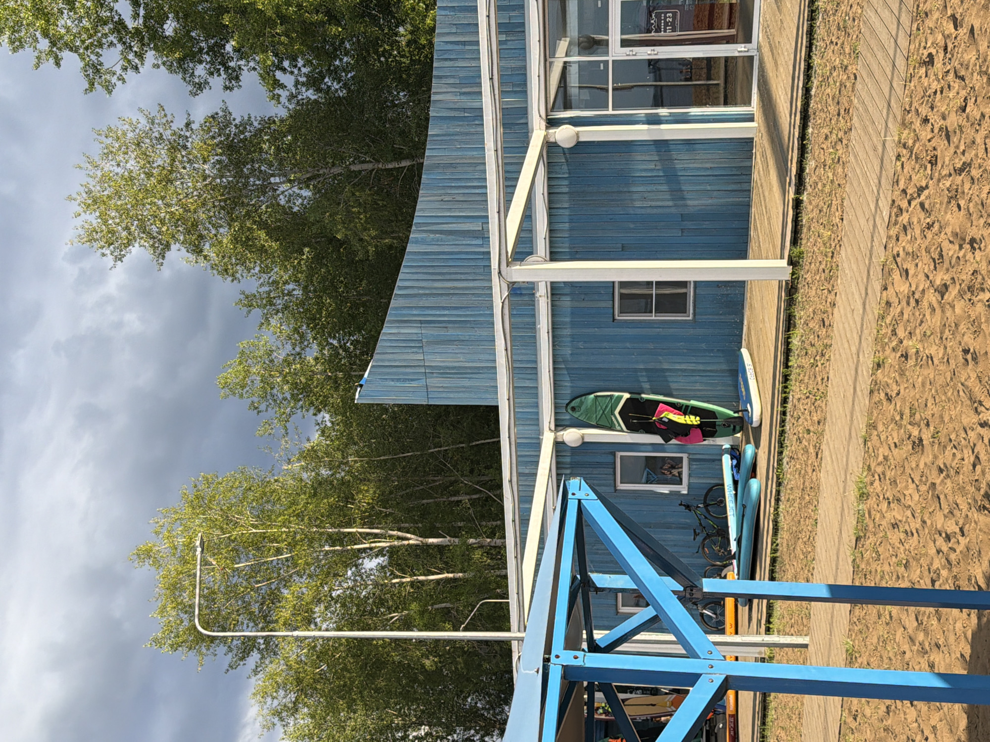A wooden beachside building painted blue with a curved roofline is seen under a cloudy sky. A white pergola extends from the front of the building, supported by white posts. Several surfboards, paddleboards, and two bicycles are arranged on the wooden deck and sand in front of the building. The building has windows and a glass door. The ground in front is sandy with a wooden walkway. The scene is likely located at a watersports rental or club on the shore of the Kama River in Chaikovsky, Russia, given the presence of watersports equipment and the typical aesthetic of such establishments in the region.