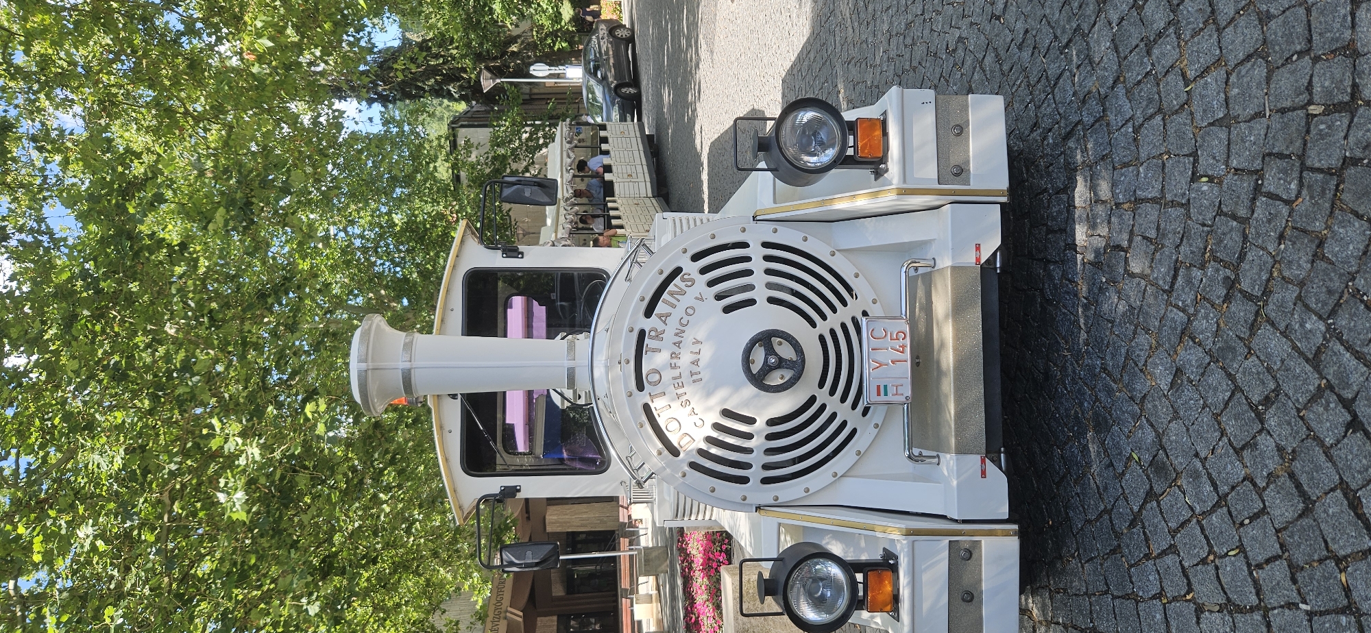 A white, toy-like train is parked on a cobblestone street in Hévíz, Hungary. The train has a large, circular front with a fan-like design and "DOTTO TRAINS CASTELFRANCO V. ITALY" inscribed on it. It has a single headlight on each side with orange turn signals below them. A license plate reading "YIC 145" is mounted on the front. Behind the engine, several passenger cars are visible, with people sitting inside. The train is surrounded by trees with lush green leaves and a clear blue sky. In the background, there are buildings and a dark gray car. A pink flowerbed is located to the left of the train.