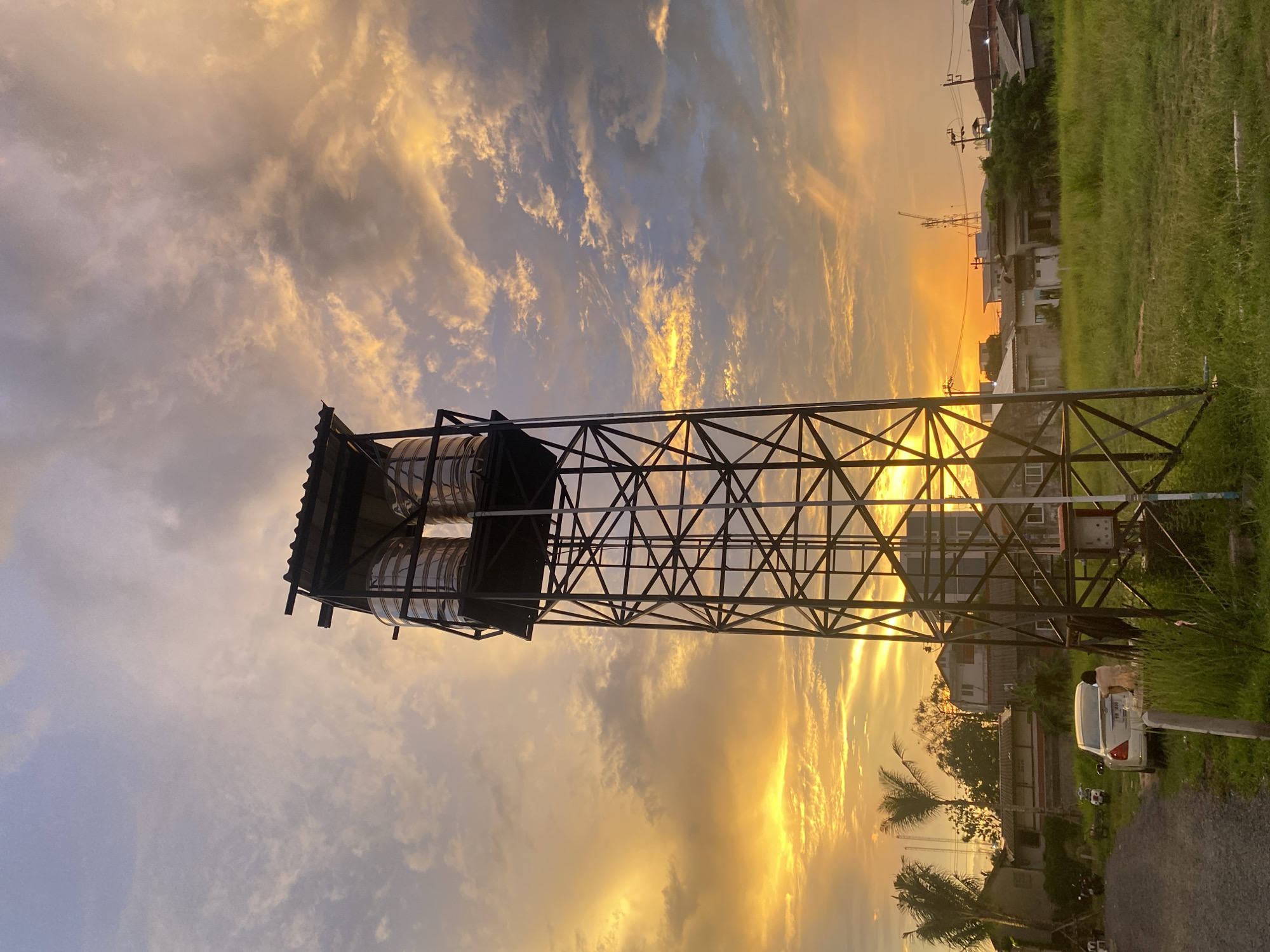 The image depicts a water tower in Choeng Thale, Thailand at sunset. The tower is constructed of a metal frame holding two cylindrical tanks under a roof. The sky is filled with clouds illuminated by the setting sun, with hues of gold and blue. In the background, houses and power lines are visible. At the base of the tower, there is a grassy area and a parked white vehicle. The scene suggests a quiet, residential area at dusk.