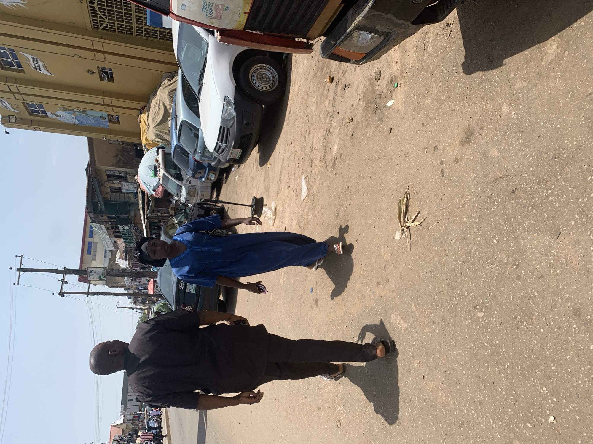 In the city of Jos, Nigeria, two people walk away from the camera on a sun-drenched, dusty street. The man on the left, with a bald head, wears a black short-sleeved shirt and black trousers, his shadow cast long on the ground. A woman on the right wears a blue dress and a dark headscarf. She holds a smartphone in her right hand and walks with a slight limp, her shadow also stretching before her. Behind them, a dark sedan is parked. Further down the street, a yellow building with "MALTINA" signage stands tall, with other structures and parked vehicles visible. The ground is a mix of packed dirt and asphalt, with scattered debris and dry vegetation. A large red and yellow truck is partially visible on the right side of the frame, its front bumper and headlight in view. The sky is clear and blue.