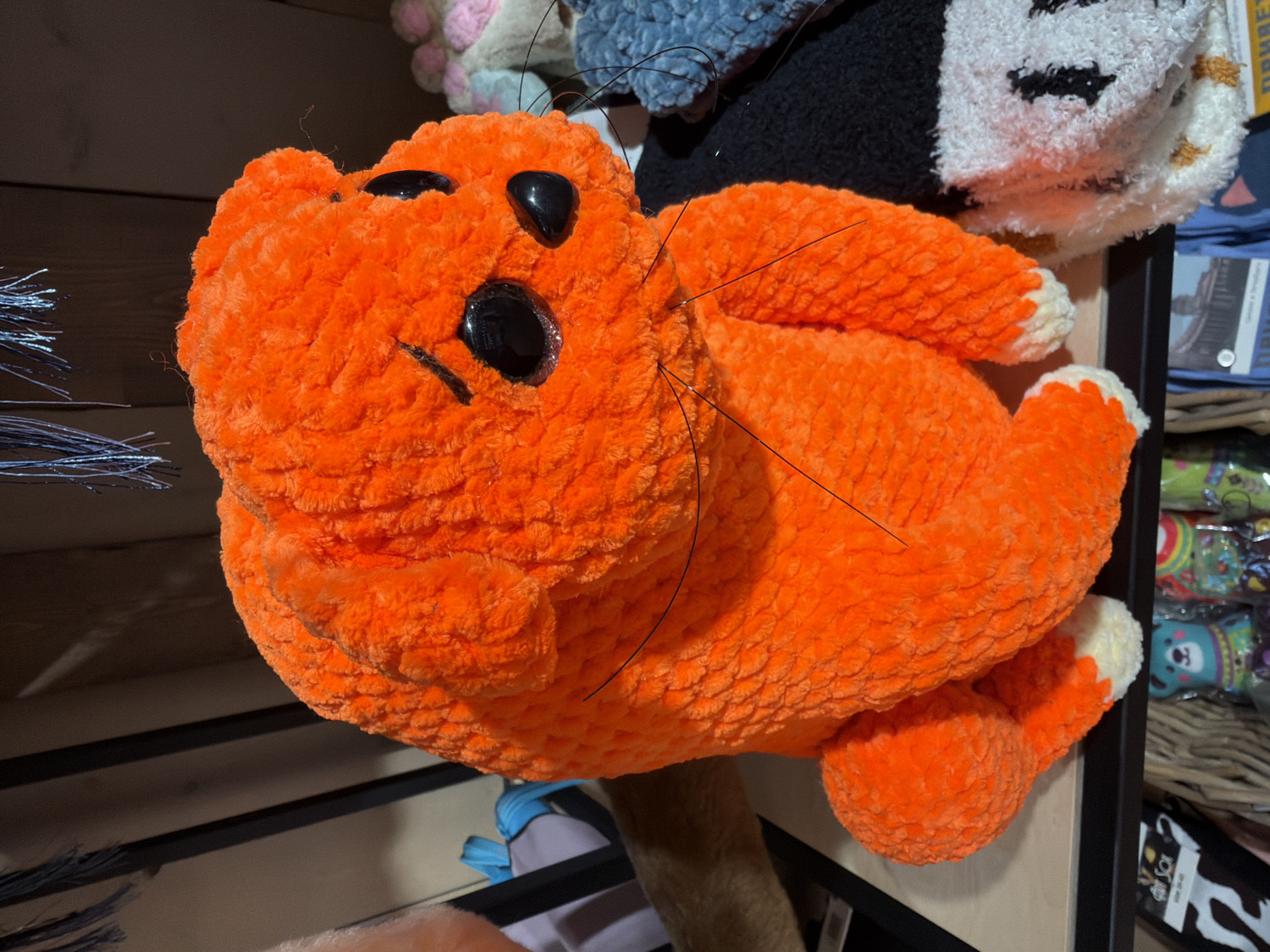 The image displays a large, vibrant orange plush toy, crocheted or knitted from chunky yarn, resembling a cat. The plush is positioned horizontally, with its head turned slightly to the left, prominently featuring its face. It has shiny black button eyes, a simple stitched black mouth, and several long black whiskers. The cat's paws are tipped with white.
The setting appears to be an indoor retail environment, likely a shop or market stall in Saint Petersburg, Russia, given the context. The background is filled with various other merchandise displayed on shelves. To the top right, parts of other plush toys are visible, including a black and white striped one and a blue or grey textured one. Below these, shelves hold a variety of products, some individually wrapped in clear plastic, showing colorful designs such as a cartoon animal face on a green package. A black shelf edge or frame runs across the mid-ground.
No specific activity is taking place; the items are static and arranged for display. The lighting suggests a well-lit interior. While most text in the background is blurry, a yellow package in the top right area partially shows what appears to be Cyrillic text, possibly "ПРИВЕТ" (Privet, meaning "Hello"), which aligns with the provided location context of Russia. A small black and white stylized cat logo is also visible on a package at the bottom right. The overall impression is one of a whimsical and colorful product display.