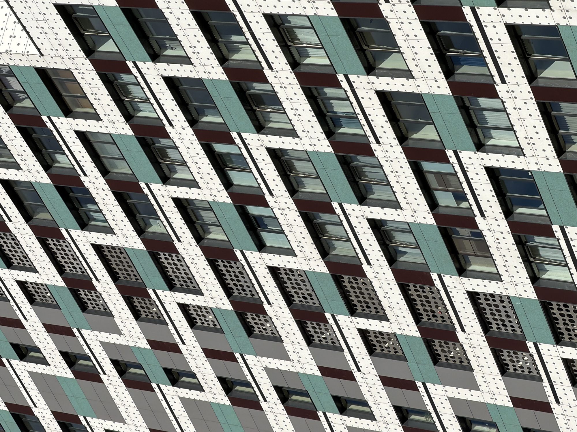 The image is a close-up, abstract view of a modern building's facade. The primary subjects are the geometric patterns and repetitive architectural elements that create a visually striking composition. We see sections of the building's exterior, characterized by a diagonal grid pattern formed by alternating white panels with dark dots, and darker colored panels, some of which are a teal green, and others a deep reddish-brown. Within this grid, there are numerous windows. Some windows are clear, revealing the interior of the building, while others are covered by perforated metal screens, suggesting ventilation or decorative elements. The windows themselves appear to be rectangular and are set within dark frames. The lighting suggests it is daytime, with shadows cast by the architectural features, indicating sunlight hitting the building. There are no discernible people or specific objects that draw attention, the focus is purely on the building's design. The overall impression is one of abstract geometry and a clean, contemporary aesthetic. The location is not identifiable from the image itself, and no text is visible.