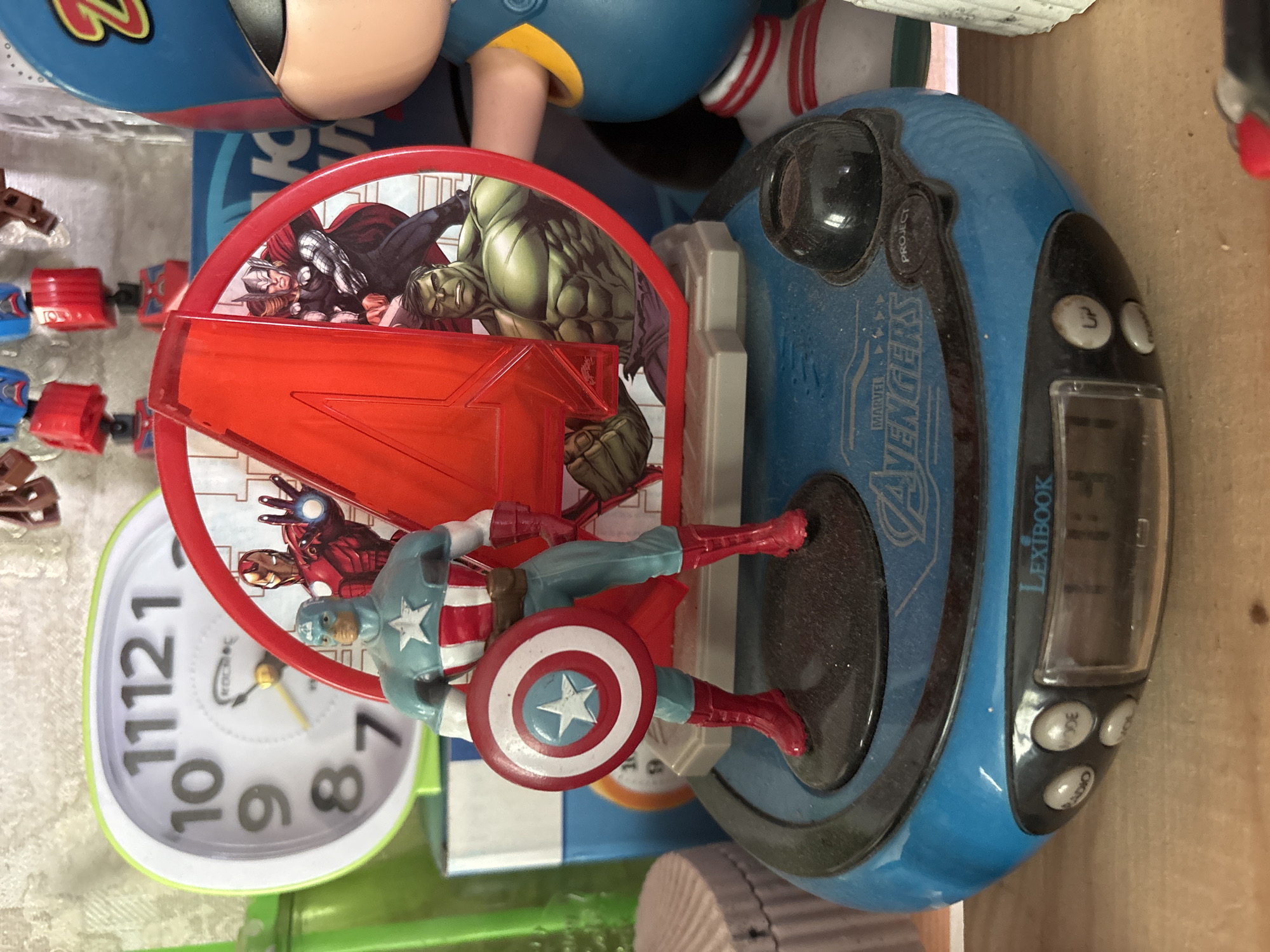An Avengers-themed digital alarm clock with a Captain America figurine on top is displayed on a wooden shelf. The clock is blue and oval-shaped, with the Marvel Avengers logo printed on its surface. The Captain America figurine is a detailed replica, holding his iconic shield. Behind the clock, an alarm clock with a lime green border is visible, showing the time 11:12. The scene appears to be set indoors, likely in a domestic environment. There is no indication that this scene is located in Sochi, Russia.