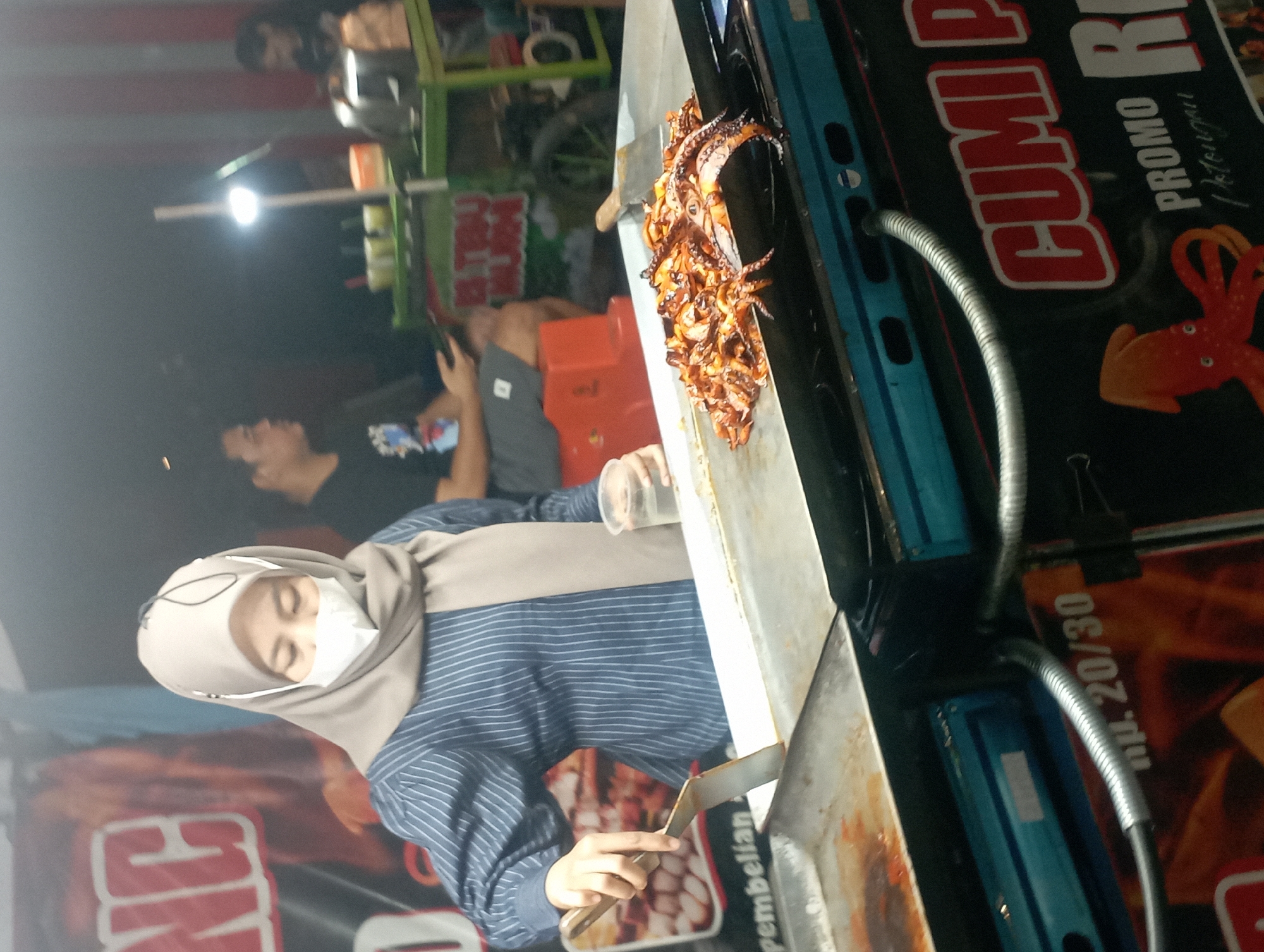 The image depicts an outdoor night scene at a food stall in Nganjuk, Indonesia, likely on a street or market. In the foreground, a woman wearing a hijab and a face mask is preparing food on a flat-top grill. She is holding a spatula in her right hand and a clear plastic cup of liquid in her left. A large pile of grilled squid is spread across the hot surface of the grill. The stall has a sign with the words "CUMI P" and "PROMO" visible, suggesting it sells squid. The background shows other people, some sitting and others standing, and more stalls or structures, illuminated by artificial lighting, creating a dimly lit and somewhat hazy atmosphere typical of night markets. The overall impression is that of a bustling, casual food establishment.