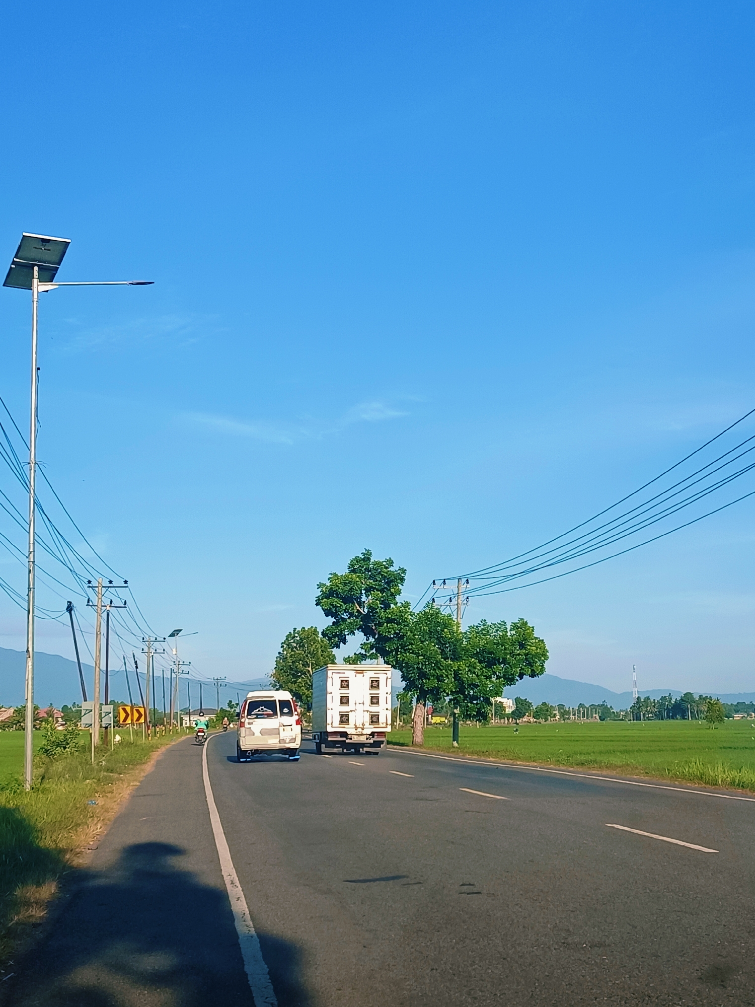 The image depicts a rural road in Kampung Jumpa, Indonesia, under a clear blue sky. The road is asphalt and features a white dividing line. On the left side of the road, there is a grassy shoulder with vegetation and a shadow of a vehicle, possibly a motorcycle. Power lines supported by wooden poles stretch across the scene, and a solar-powered street light is visible. A yellow directional sign with black arrows is also present on the left.
In the middle of the road, a white van is traveling away from the viewer. To the right of the van, a white truck with a box trailer is also moving in the same direction. Further down the road, a person on a motorcycle is visible.
On the right side of the road, a lush green rice field stretches into the distance, bordered by trees. In the background, mountains are visible, and a tall radio tower stands on the horizon. The overall impression is a bright, sunny day in a tranquil agricultural setting.
