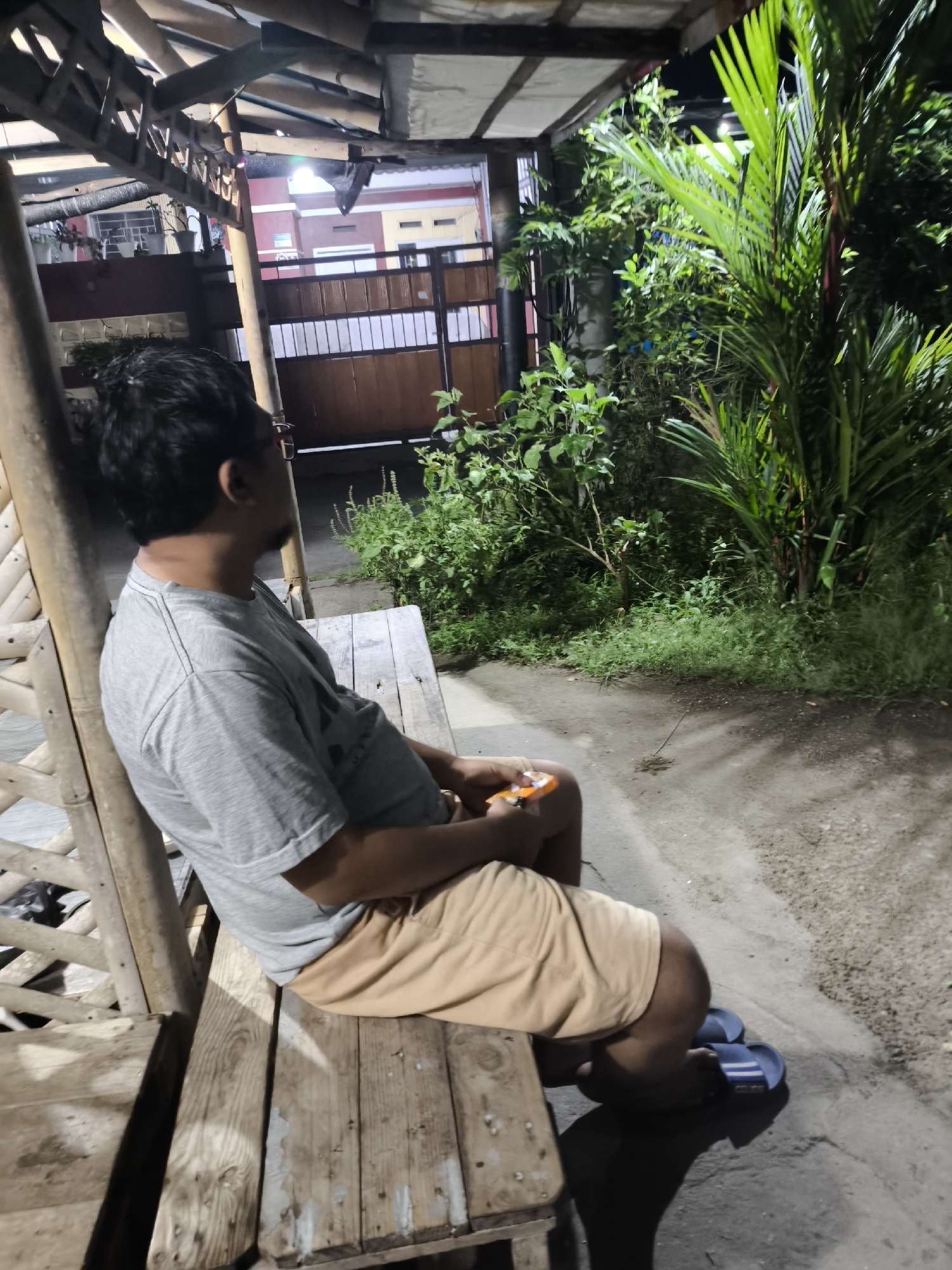 A man is sitting on a wooden bench under a covered structure with a bamboo-like railing. He is wearing a grey t-shirt and tan shorts. The man is holding a small orange object in his left hand. In the background, there are lush green plants, a house with a red and white facade, and a fence. The scene appears to be outdoors at night, illuminated by artificial lighting. The location is stated to be Cilegon, Indonesia.