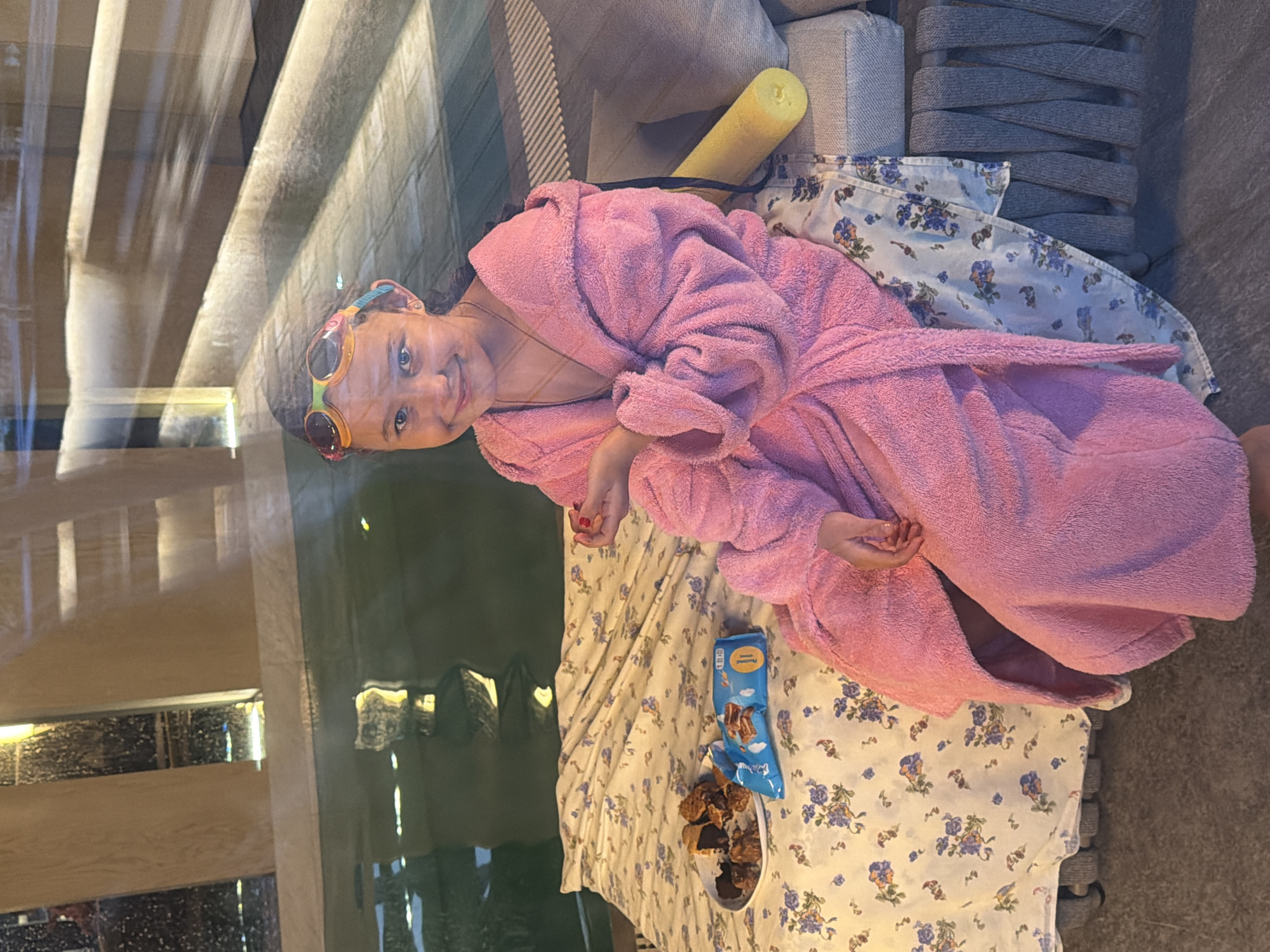 A young girl, approximately 8 years old, is lying on a bed covered with a white floral print sheet. She is wearing a pink terrycloth robe and colorful sunglasses. A yellow pool noodle lies beside her. Next to her are a plate of cookies and an open package of cookies. The background is blurred, but shows what appears to be an interior with a staircase and a reflection of an outdoor scene with possible vegetation. The image was taken through a window. There are no indications of the location being Shamoni, Russia.