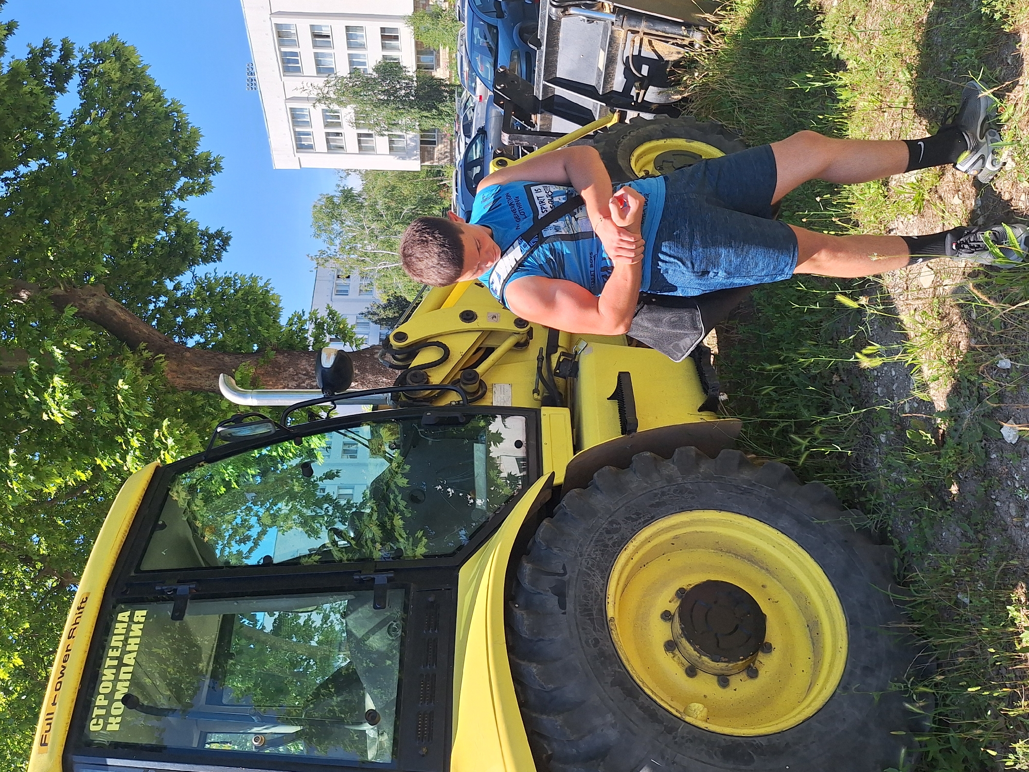 In Varna, Bulgaria, a young boy stands next to a yellow tractor under a leafy tree. The boy, wearing a blue sleeveless shirt and blue shorts, is looking at his wrist. He has a black bag slung over his shoulder. The tractor has "Full-power Shift" and "СТРОИТЕЛНА КОМПАНИЯ" (Construction Company) written on it. In the background, there are buildings and parked cars. The environment is grassy and appears to be a sunny day.
