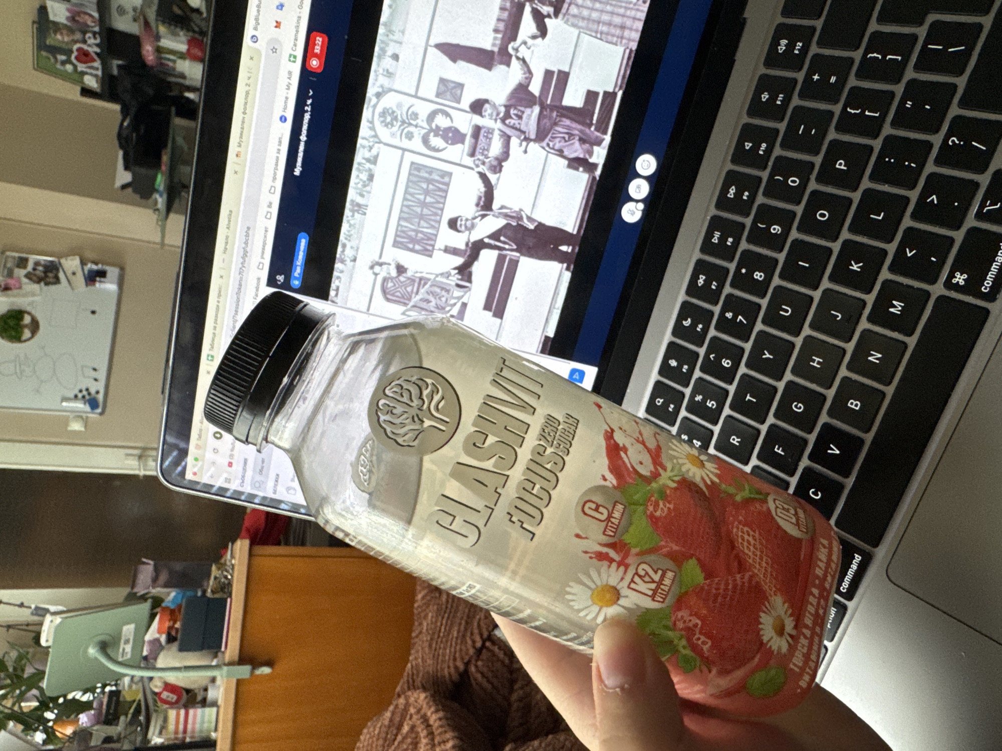 Here is a description of the image:
**Scene:** A close-up shot inside an indoor setting, likely a home office or study area.
**Subjects:** A hand is holding a bottle of "CLASHVIT Focus Zero Sugar" flavored with forest strawberry and chamomile. The bottle is mostly visible.
**Objects:** A laptop is open in the background, displaying a video or image on the screen. A keyboard is next to the bottle. A desk is visible behind the laptop.
**Environment:** A room with a cluttered desk and various items. A white board is on the wall.
**City (Implied):** There is not enough information to determine the city. Bulgaria is only implied by the drink.