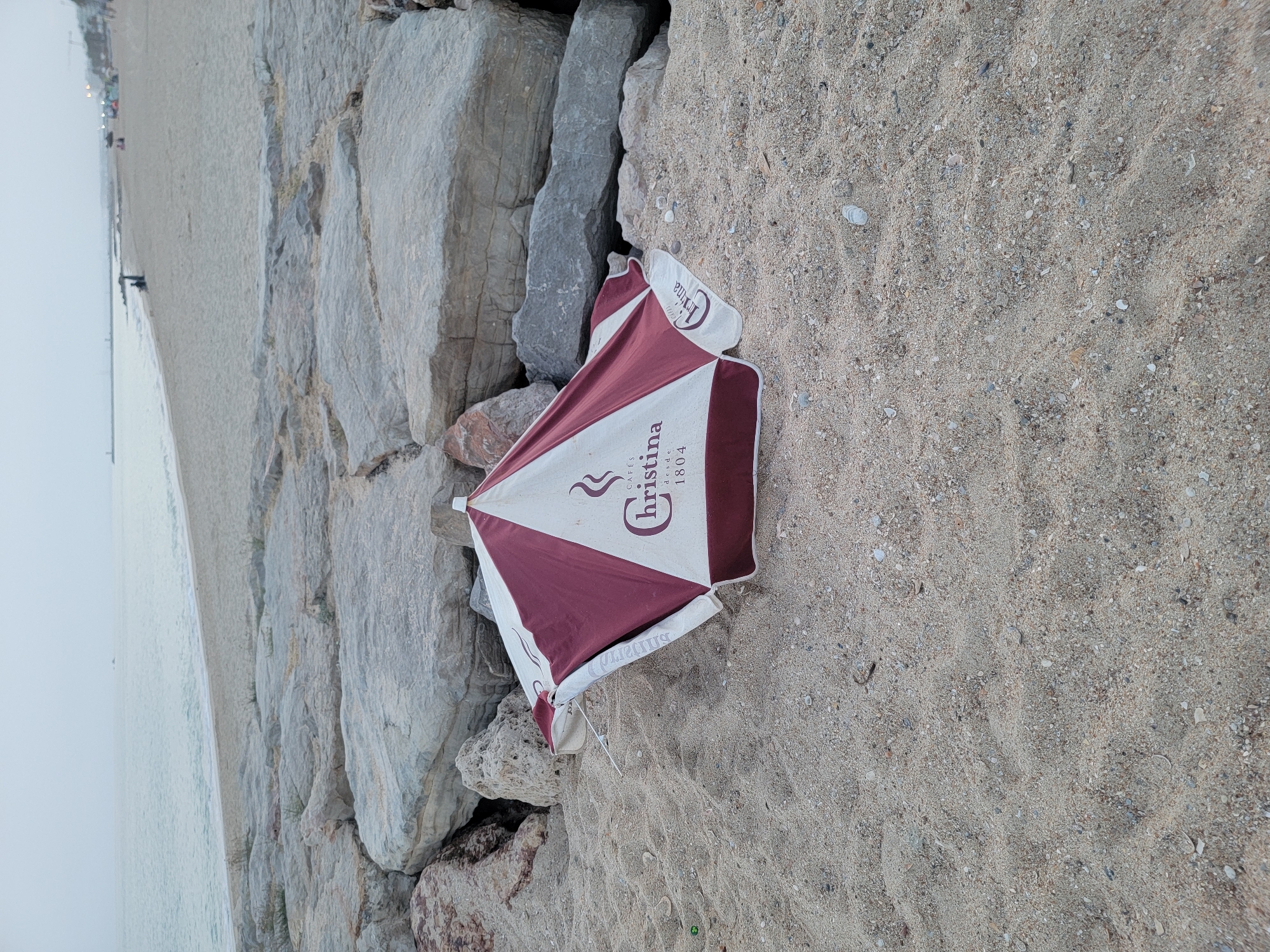 A collapsed beach umbrella lies on the sand in Quarteira, Portugal, near a wall of large, grey rocks. The umbrella is primarily white with maroon geometric patterns. The words "CAFE Christina desde 1804" are visible on a section of the umbrella. The sand is coarse with small shells and debris. In the background, the ocean meets a sandy beach, with a breakwater and distant figures visible. The sky is overcast and the overall lighting suggests it is either early morning or late evening.
