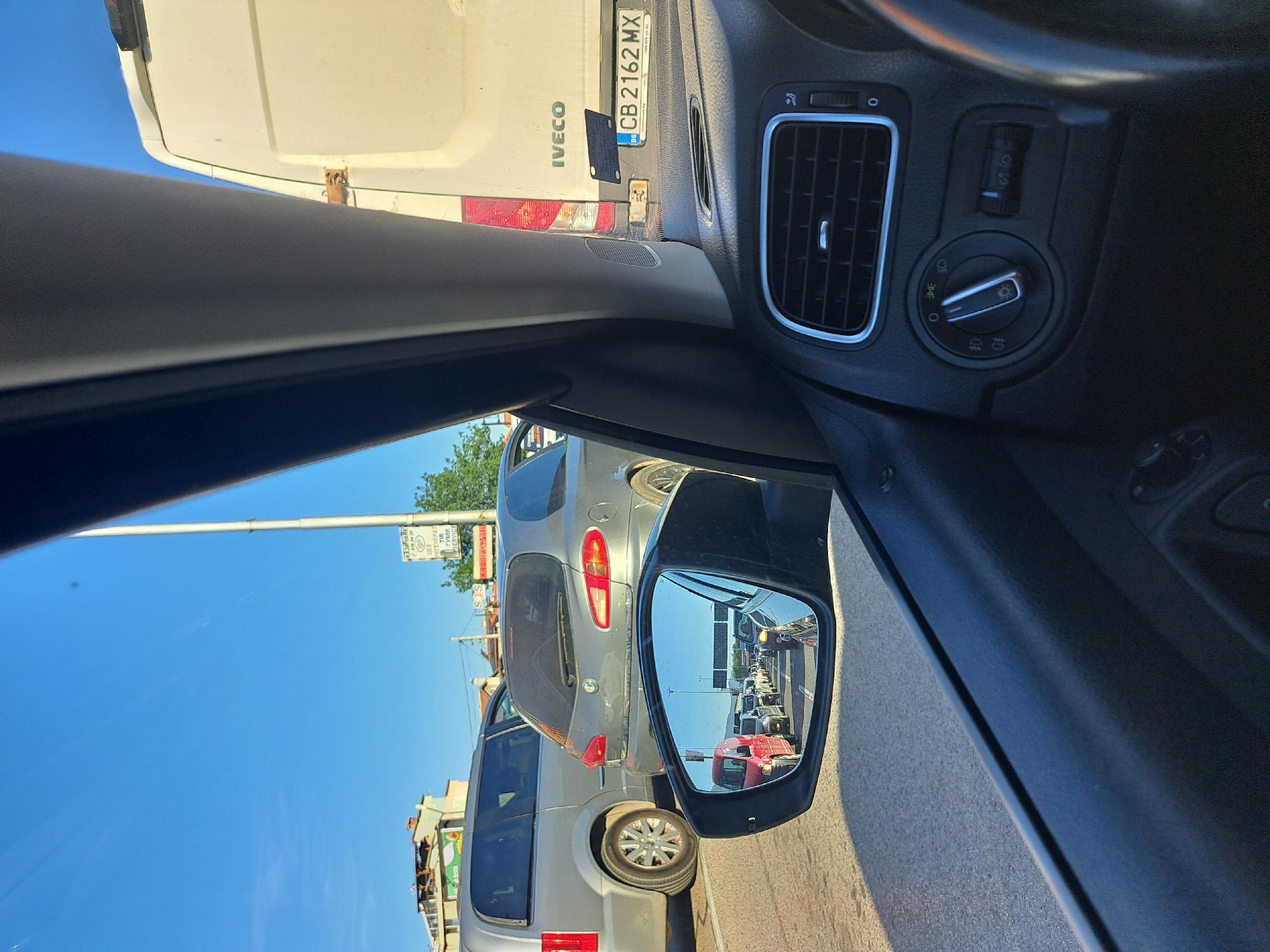 An eye-level, medium shot captures the interior of a car looking out at a traffic jam under a clear blue sky. The driver's side door panel and a portion of the dashboard, including the air vent and light controls, are visible in the foreground. In the middle ground, a silver Alfa Romeo is parked to the right of a silver minivan, with both vehicles facing away from the viewer. A side-view mirror on the driver's side reflects a long line of cars stretching into the distance, suggesting heavy traffic. In the background, buildings and trees are visible, as well as a white Iveco van with a Bulgarian license plate reading "CB 2162 MX." The scene appears to be in a city setting, possibly during daytime, given the bright sunlight and clear sky.