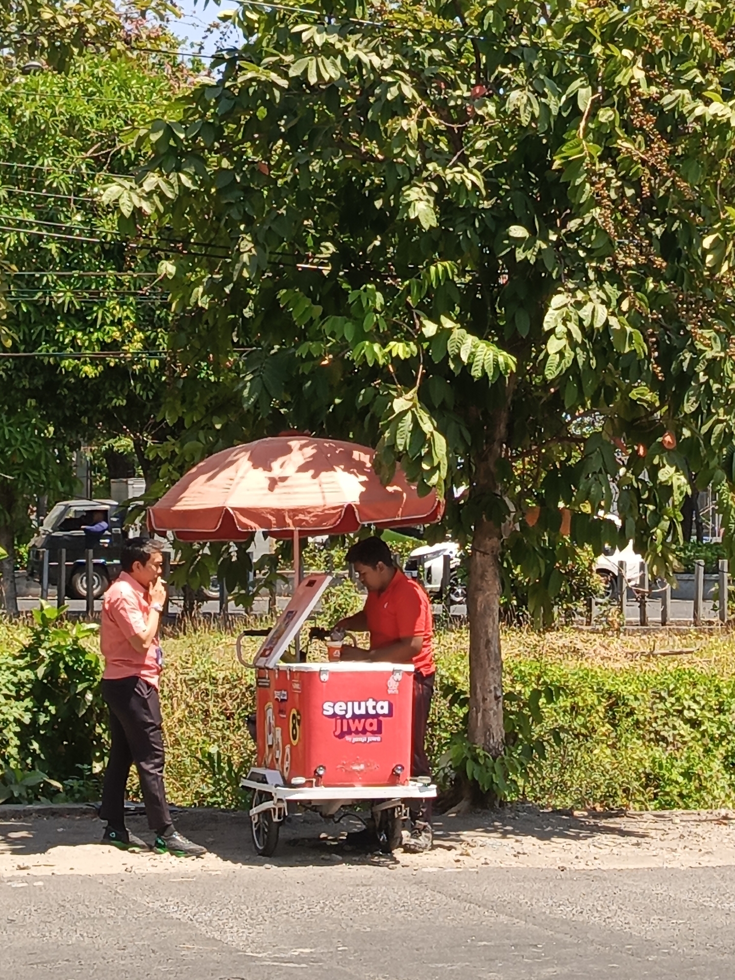 A man in a salmon-colored shirt and black pants stands to the left of a red ice cream cart, smoking. He is looking towards the cart, where another man in a red shirt is serving an order. The cart is under a large tree with green leaves and some reddish-brown fruit or berries. The scene appears to be outdoors, possibly on a roadside in a city, with vegetation and a fence in the background. A dark-colored vehicle is parked behind the man in the salmon shirt. The text on the cart reads "SEJUTA JIWA" in white letters with a pink outline. The ground is paved, suggesting an urban setting.