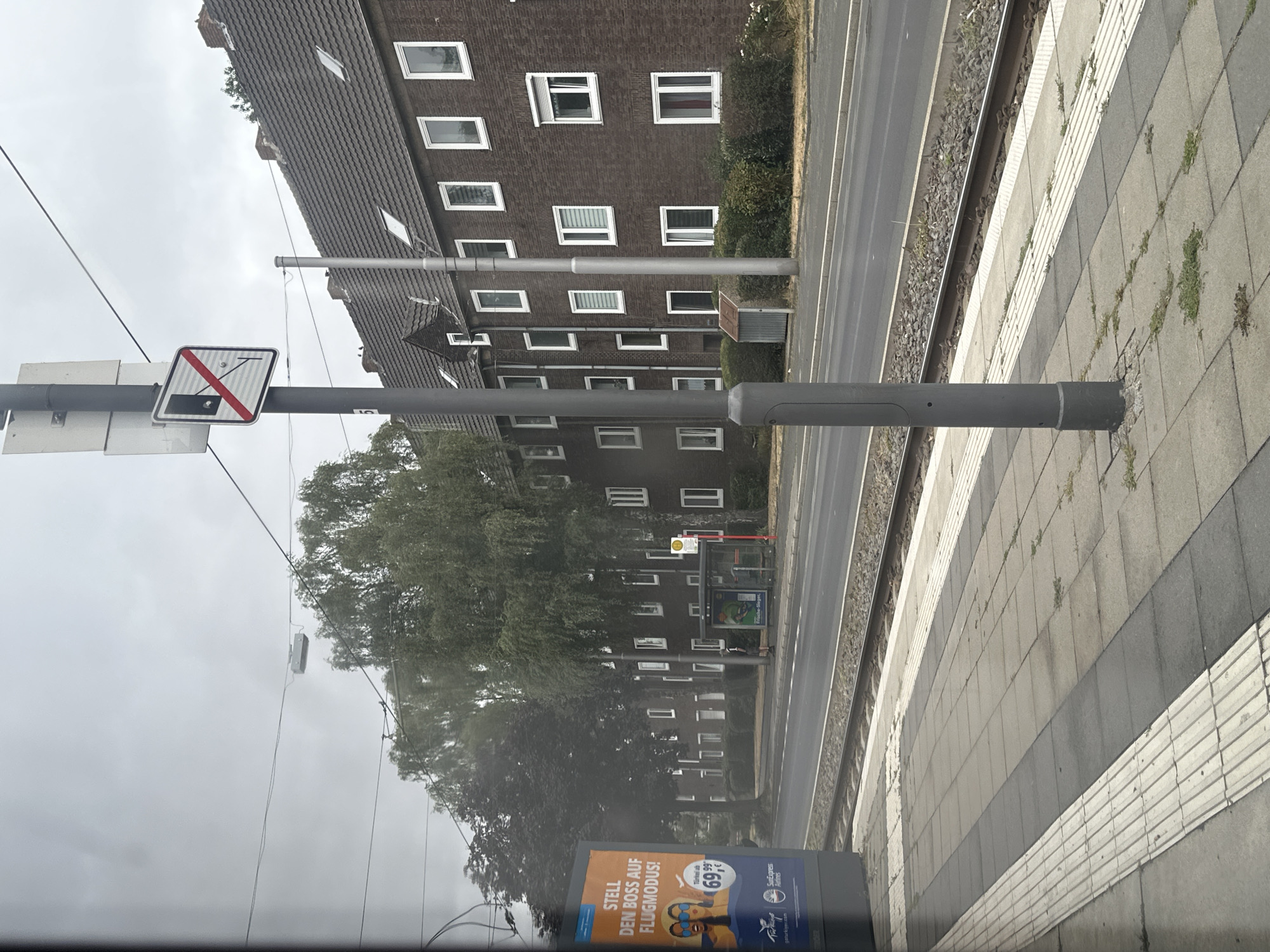 This image captures a view from a tram platform in Krefeld, Germany. The foreground shows a grey tiled platform with tactile paving in white. To the left, a large advertising board displays a travel advertisement with an image of two women wearing sunglasses, text in German including "STELL DEN BOSS AUF FLUGMODUS!" and pricing information, along with logos for "goturkiye.com" and "SunExpress Airlines". A tram track runs alongside the platform.
In the mid-ground, a street runs parallel to the tram tracks. On the other side of the street, a row of attached brick apartment buildings with dark tiled roofs and white-framed windows are visible. Some of the windows have white roller shutters or blinds. A small red and yellow bus stop shelter with a white roof stands on the sidewalk in front of the buildings, with a sign visible. A person is standing near the bus stop.
The background is dominated by a canopy of lush green trees, obscuring a cloudy, overcast sky. A grey pole with traffic signs, including a regulatory sign with a diagonal red line over a black rectangle and a circular element, stands prominently on the platform. Overhead, electrical wires for the tram system are suspended across the frame. The overall scene depicts a typical urban streetscape with public transportation infrastructure and residential buildings on a cloudy day.