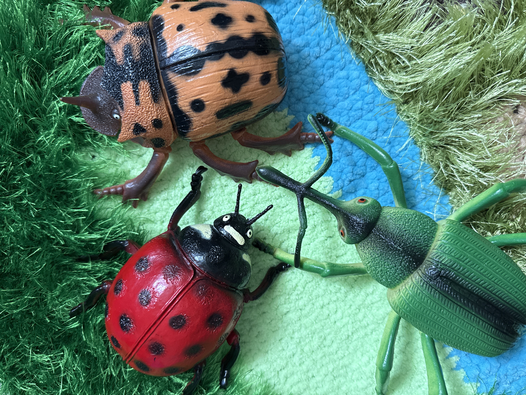 This media file displays a close-up, top-down view of three toy insects arranged on a textured surface. The subjects are two beetles and one ladybug, all rendered in plastic with a toy-like appearance.
The largest insect, positioned in the upper left quadrant, is a brown and black spotted beetle. It has a prominent horn on its head and several articulated legs. To the lower left is a red ladybug with black spots, also featuring visible legs and antennae. On the right side of the image is a bright green beetle with a long, slender snout. Its legs are also visible, as are its red eyes and segmented body.
The insects are situated on a background that appears to be a textured mat or fabric. There are patches of green, fuzzy material resembling grass, interspersed with areas of blue and light green textured fabric. The overall impression is of a miniature outdoor scene, perhaps a playful arrangement.
No specific activity is depicted; the insects are stationary. Notable details include the glossy finish on the ladybug and the matte texture on the green beetle's body. The lighting suggests it is daytime, with no indication of weather conditions. The location context of Vladivostok, Russia, is not visually represented in the image. There is no visible text in the media file.