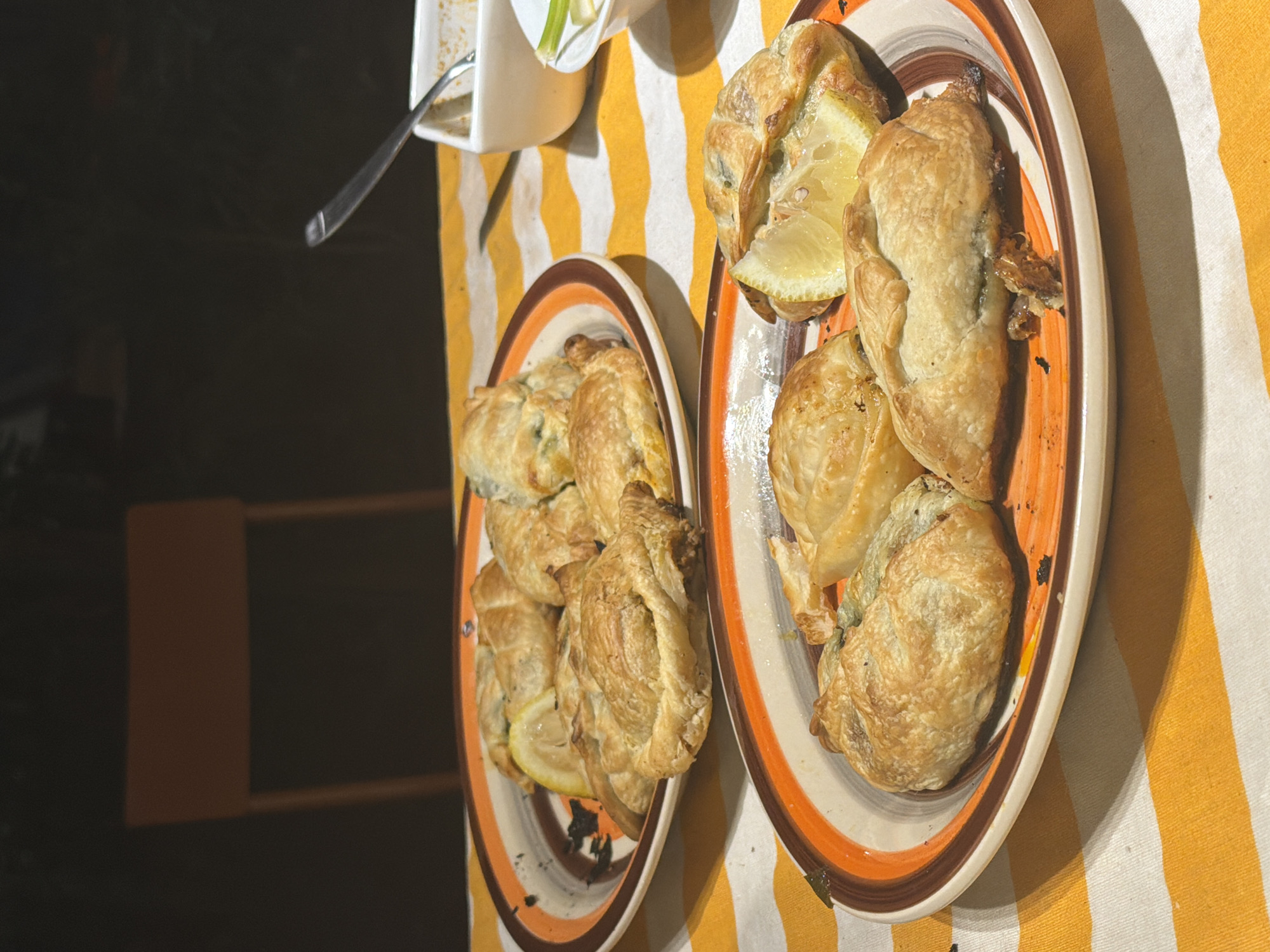 Two plates of pastries with lemon wedges are arranged on a yellow and white striped tablecloth. A white container with silverware is also visible on the tablecloth. The scene appears to be indoors, possibly in Buenos Aires, Argentina, but there are no specific indicators to confirm this location. The focus is on the pastries and their presentation. There are no people visible in the frame.