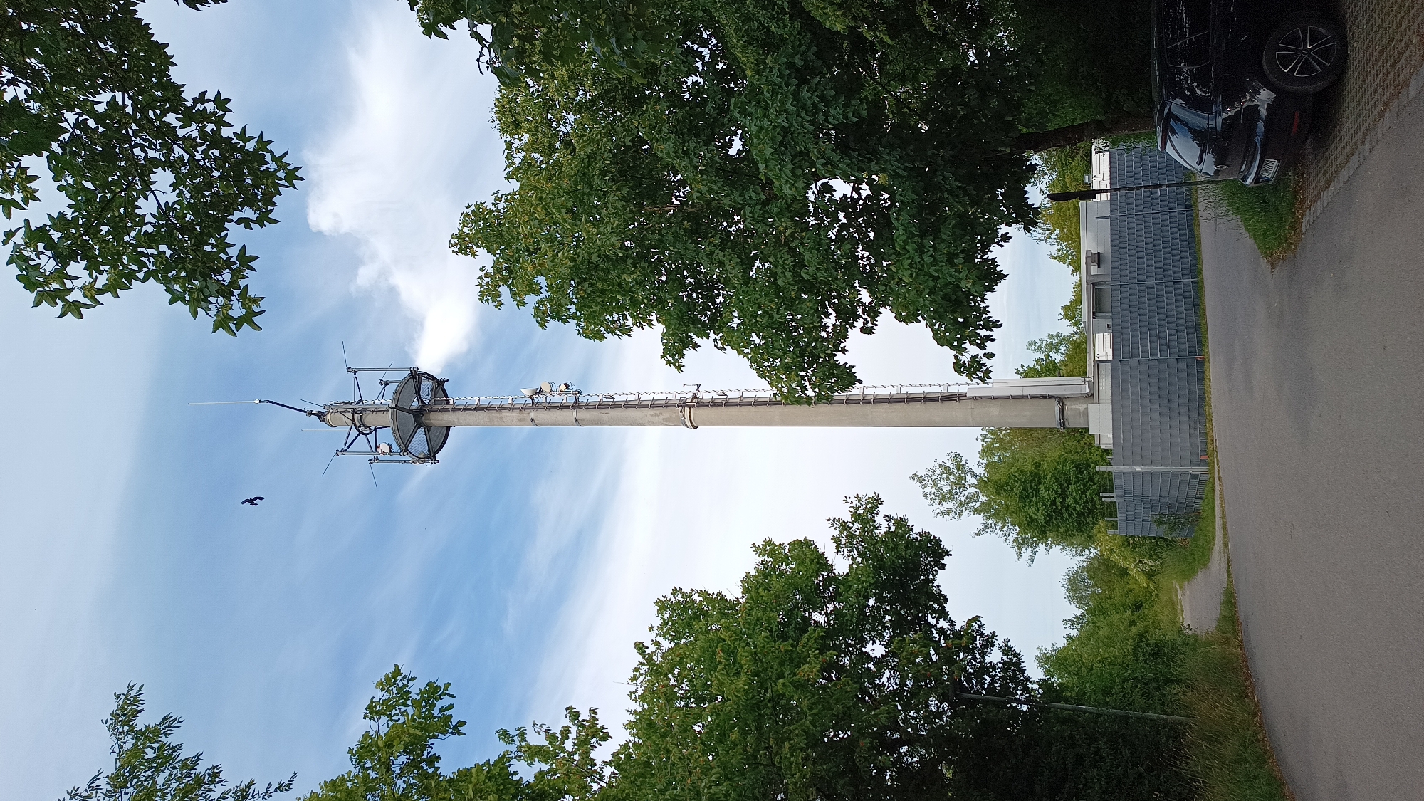 A concrete telecommunications tower stands tall against a cloudy sky, flanked by lush green trees. A narrow, paved path leads away from the foreground into a wooded area. A black car is parked on the right side of the frame, next to a gravel area. The base of the tower is enclosed by a grey metal fence, behind which are several utilitarian buildings. The scene appears to be located in Unterhaching, Germany. There are no people visible in the image.