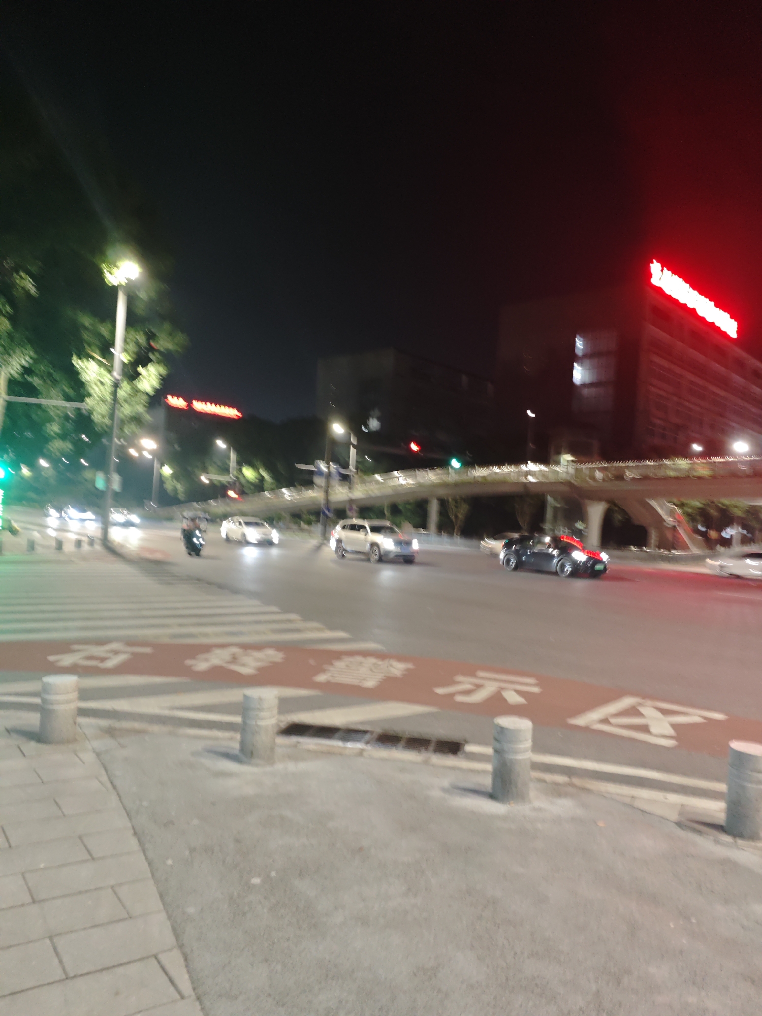 At night in the Chongqing High-tech Zone, China, a busy intersection is captured. A crosswalk with red markings and white stripes is prominently featured in the foreground. Several cars, including a black sports car and a white sedan, are visible on the road, some of which are in motion. A scooter with a rider is also present. In the background, a pedestrian bridge spans across the road, illuminated by lights. Tall buildings are visible, with a large red neon sign on one of them. Streetlights are scattered throughout the scene, casting a glow on the environment. The overall atmosphere suggests a bustling urban setting at night.
