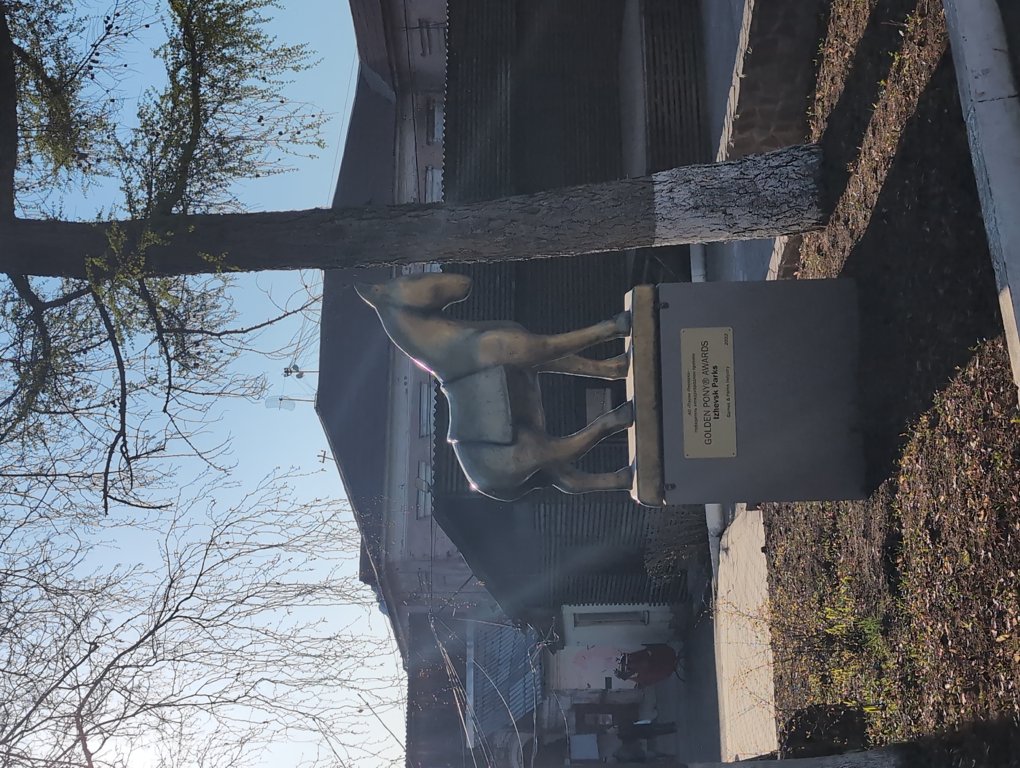 The image shows a bronze statue of a pony on a concrete pedestal. The pedestal has a metal plaque with the inscription "GOLDEN PONY® AWARDS Izhevsk Parks" in Russian and English. The statue is positioned next to a tree with a partially painted trunk. The background shows a building with a corrugated metal facade. Bare branches are visible in the lower left corner. The location is most likely in Izhevsk, Russia.