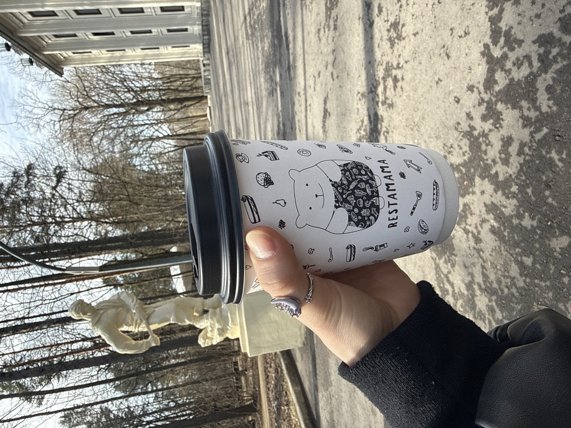A hand holding a paper cup with the text "RESTAMAMA" is visible. The hand belongs to a person wearing a black sweater or jacket and a silver ring. The background consists of a statue, trees, and a building. The location is identified as Moscow, Russia.