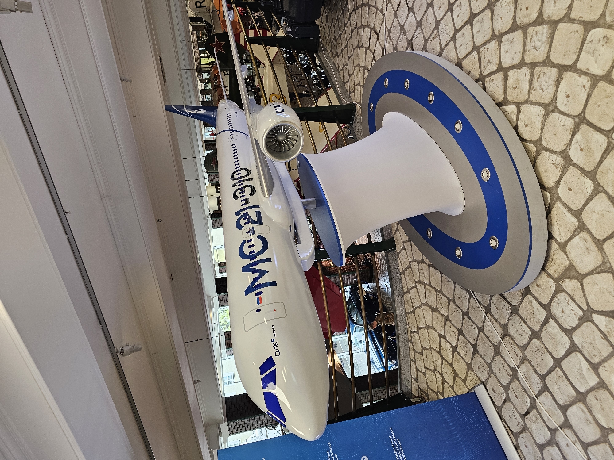 The image shows a model of an MC-21-310 aircraft in what appears to be a museum or exhibition hall. The aircraft is painted white with blue accents and markings. The aircraft model is suspended above a white stand with a circular grey and blue base. The floor is paved with light-colored stones. In the background, there is metal railing and other museum exhibits are visible. The location is likely in or near Архангельское, Russia, given the Cyrillic markings on the aircraft.