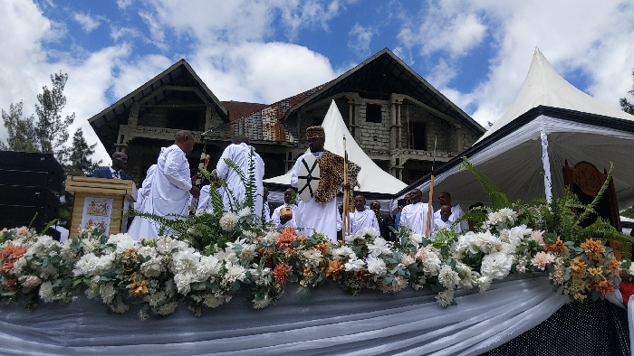 The image shows a group of people, primarily men, dressed in white robes, standing behind a white table adorned with a large floral arrangement. Several of the men hold various objects, including a shield, spears, and a cross. The event is being held outdoors, with a large white tent, a stone building with a wooden roof, and lush greenery visible in the background. The sky is partly cloudy and blue. The location is Rukoko, Democratic Republic of the Congo, at coordinates -1.624, 29.258. The scene is reminiscent of a religious ceremony. The men in the foreground appear to be participating in a ritual or procession, while others are watching and engaging in conversations.