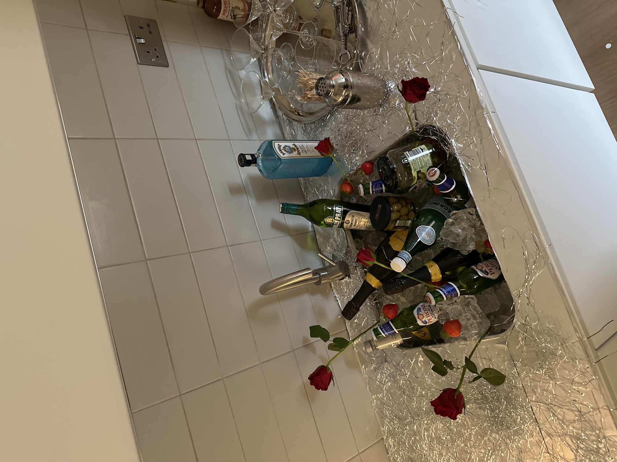 The image depicts a modern interior setting, likely a bathroom or kitchenette, with a tiled wall featuring an electrical outlet. A sink filled with ice, several bottles of alcoholic beverages including various types of wine and a bottle of Bombay Sapphire gin, as well as bottled olives, roses, and strawberries placed on top of the ice for aesthetic appeal. Other objects include a cocktail shaker, glassware, and decorative silver tinsel-like material. The lighting is bright. No people are visible. The image does not provide enough information to determine if the location is in London, United Kingdom.