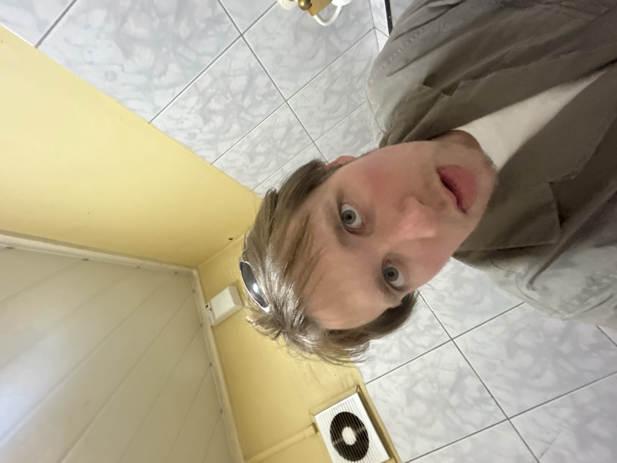 A man is taking a selfie in a small room with yellow walls and white tiled floors. The walls are lined with vertical white panels. An air conditioning unit is on the lower right corner of the wall. The man is wearing a tan jacket.