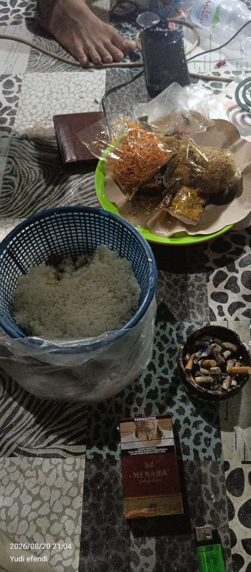 The image depicts a dimly lit, casual gathering, possibly a meal, set on a patterned surface. In the foreground, a blue plastic basket contains a generous portion of white rice. To the right of the rice, a small, dark-colored ashtray is filled with used cigarette butts. A pack of "MENARA" brand cigarettes and a green lighter are also present. In the background, on a bright green plate, are several food items wrapped in clear plastic, including what appears to be fried noodles and other savory dishes. A brown wallet lies to the left of the plate. A person's bare foot is visible at the top of the image, resting on the patterned surface. The overall impression is one of a relaxed, informal setting. The image is timestamped "2026/08/20 21:04" and attributed to "Yudi efendi," suggesting it was taken in Rawajitu Selatan, Indonesia.