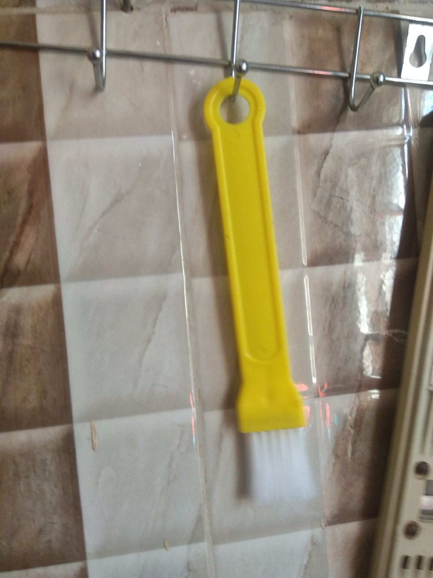 A yellow brush with a hole in the handle hangs from a metal rack attached to a tiled wall. The tiles are a pattern of light and dark brown squares. A clear plastic sheet is partially visible behind the brush. To the right of the brush, a portion of a beige electronic device is visible. The scene appears to be indoors, likely a kitchen or bathroom in Jalingo, Nigeria. There are no people or animals present.