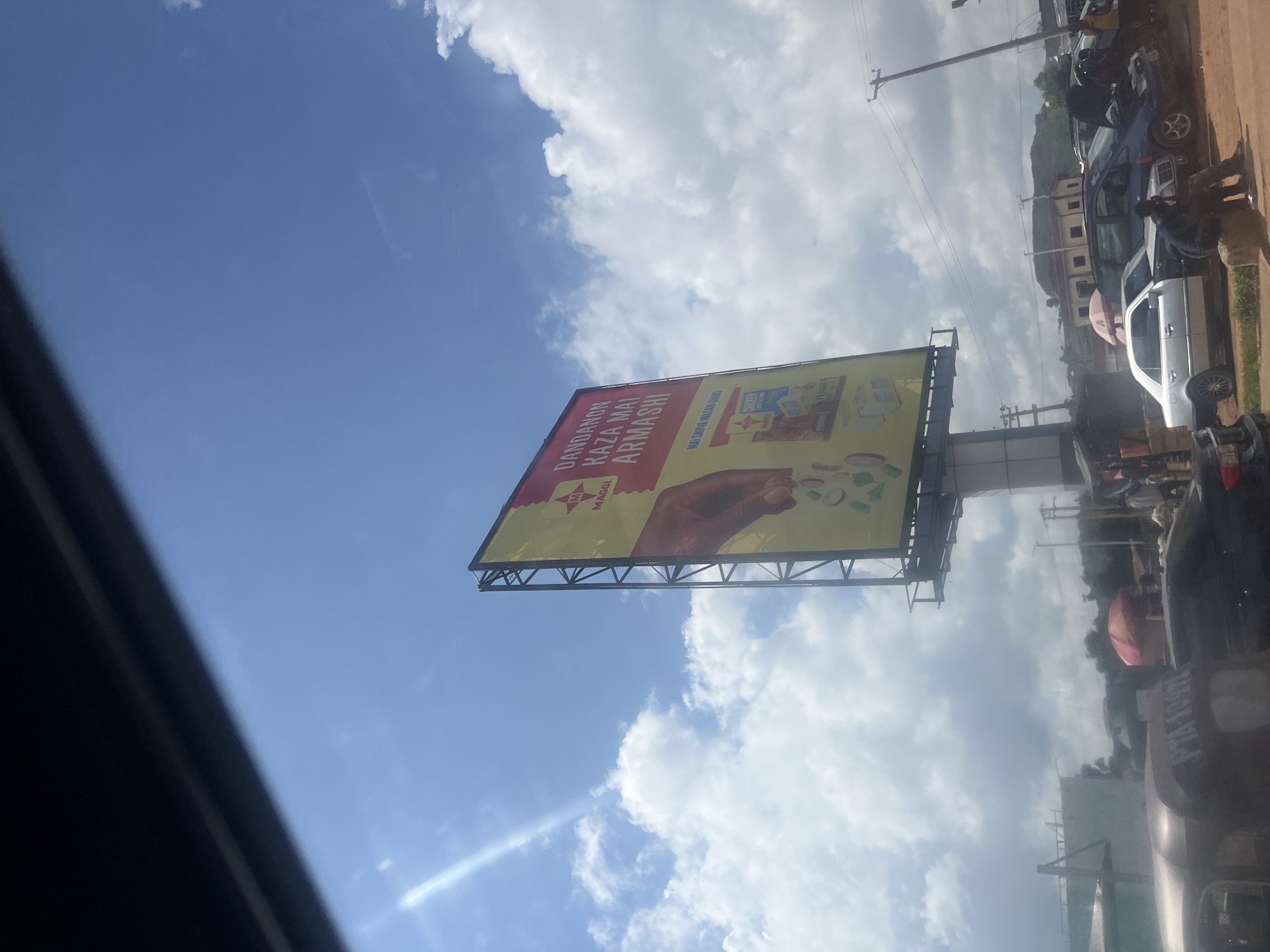 The image shows a street scene in Jos, Nigeria, dominated by a large billboard for Maggi seasoning. The billboard, predominantly yellow and red, features text in a language that appears to be Hausa, with images of Maggi products and ingredients. A human hand is prominently displayed, seemingly adding ingredients to a dish. Sunlight streams in from the left, creating lens flare and casting bright highlights on the sky and clouds.
Below the billboard, a busy street is visible with various vehicles, including cars, a minivan, and a three-wheeled transport vehicle (Keke Napep). Several people are present, some interacting with the vehicles or standing by the roadside. One individual is seated on a rock in the foreground. In the background, buildings and trees are visible, along with a hazy sky filled with scattered clouds. The overall impression is of a typical urban environment in Jos on a bright, partially cloudy day.