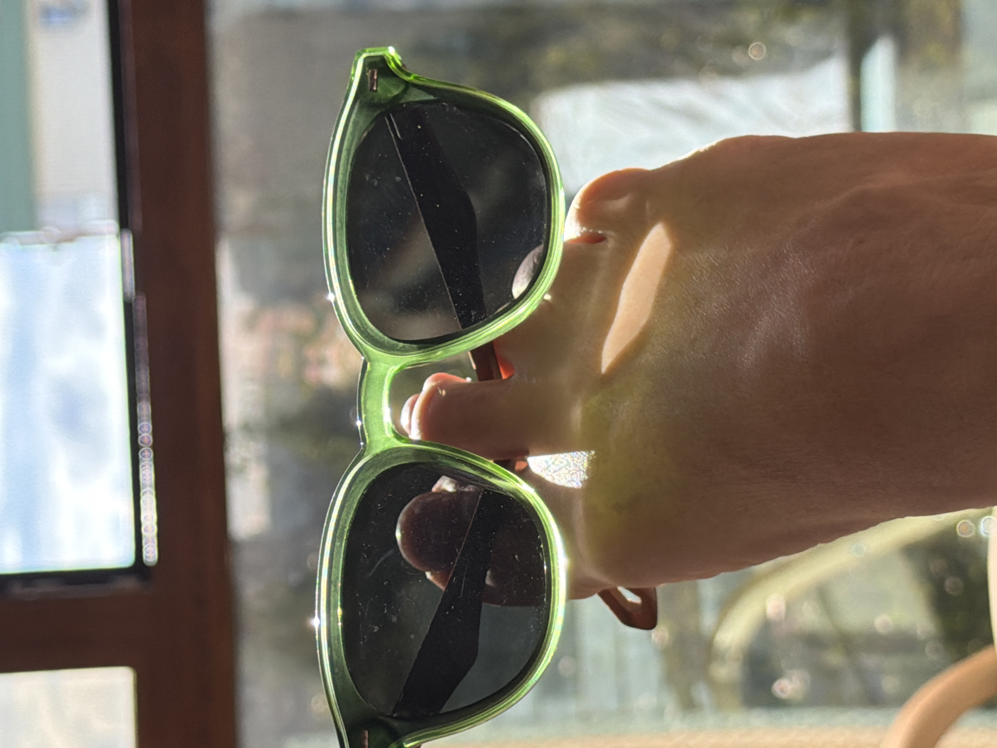 A close-up, eye-level shot shows a hand holding a pair of light green sunglasses with dark lenses in front of a blurred background. The hand is positioned to frame the sunglasses, with the thumb visible through the left lens. The background appears to be a window with sunlight streaming through, creating a bokeh effect of circular highlights. The overall scene suggests an outdoor or indoor setting near a window. The location in the city of Surgut, Russia, is not discernible from the image.