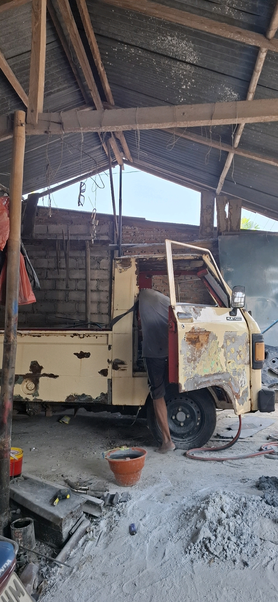 A person is working on the door of a distressed, light yellow utility truck in a makeshift, open-air garage in Desa Gagaksipat, Indonesia. The truck is heavily weathered, with significant rust and peeling paint evident on its body panels. Tools and construction materials, including a bucket of water and a pile of concrete mix, are scattered on the dusty ground. The garage has a corrugated metal roof supported by wooden beams, and the back wall appears to be constructed of brick. The overall scene suggests ongoing repair or restoration work in a rustic environment.