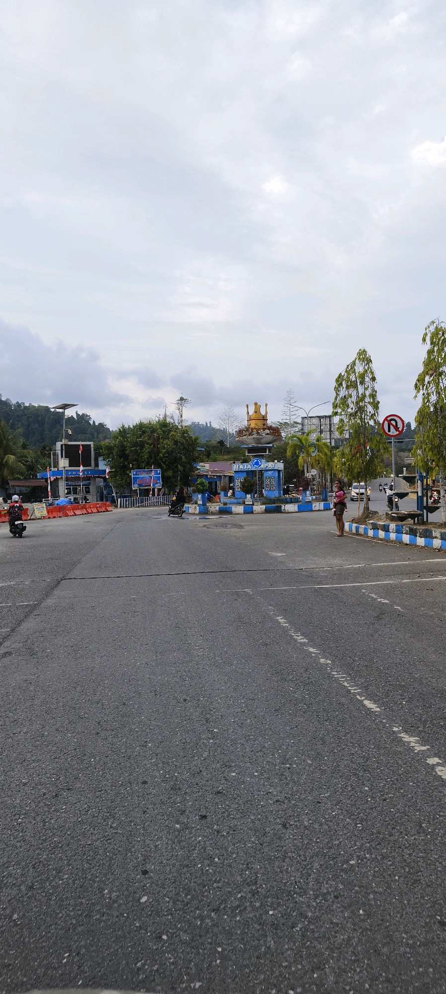 The image shows a street scene in Waisai, Indonesia. In the foreground, a paved road with white lane markings runs towards the viewer. In the middle ground, there is a roundabout with a monument topped by a golden crown. Several buildings are visible behind the roundabout, including a blue building with "POLISI" (Police) signage. Orange traffic barriers are set up on the left side of the road. Trees line the right side of the roundabout, and a "no U-turn" traffic sign is visible. A woman carrying a child is standing near the right edge of the roundabout. Several motorcycles and cars are present, with two motorcycles in motion on the left side of the road and a few vehicles in the distance. The sky is overcast with clouds. The overall environment appears to be a town or city street with some public infrastructure and traffic.