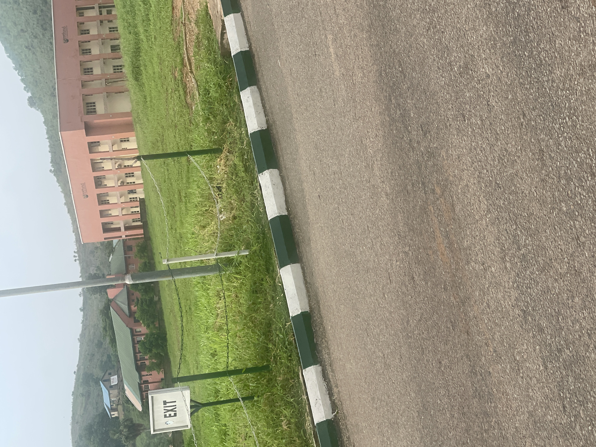 The image shows an outdoor scene with a road in the foreground and buildings and hills in the background. A paved road with a curb painted with alternating green and white stripes runs across the bottom of the frame. Behind the curb, a grassy area is visible, separated by a chain-link fence supported by green posts. A white sign with the word "EXIT" in black letters is mounted on a green post.
Further back, several buildings are visible. One is a long, pink building with multiple windows and a flat roof, appearing to be a modern educational institution or office building, with a logo that reads "redfund" on its facade. To the left, another building with a green roof and pink walls is partially visible, and behind it, a smaller building with a blue roof is situated on a hill. The landscape is characterized by rolling green hills, suggesting a semi-rural or suburban environment. The sky is overcast. The scene is likely located in Lokoja, Nigeria, given the context of the provided information. There are no people or animals clearly visible in the image.