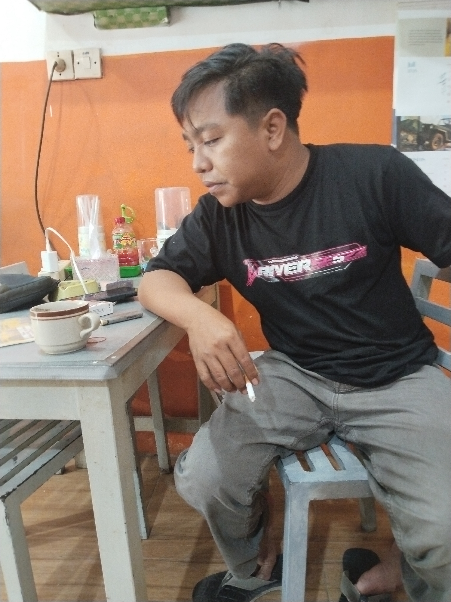 The image shows a man sitting at a table in an indoor setting. He is wearing a black t-shirt with a pink and white graphic that appears to read "RIVER" and some numbers. He is also wearing grey pants and sandals. The man is holding a lit cigarette in his right hand and looking downwards.
On the table in front of him, there is a white ceramic cup with a brown rim, a cigarette pack, and a pen. Behind the cup, there are several items including a bottle of red liquid, a clear plastic cup filled with white straws, and a clear plastic container. To the left of the table, a black bag is visible.
The wall behind the man is painted orange. There is an electrical outlet with a black cord plugged into it mounted on the wall. A calendar is also visible on the wall in the background, showing images of what appear to be vehicles. The flooring appears to be wooden planks.
The location is identified as the city of Prigi, Indonesia.