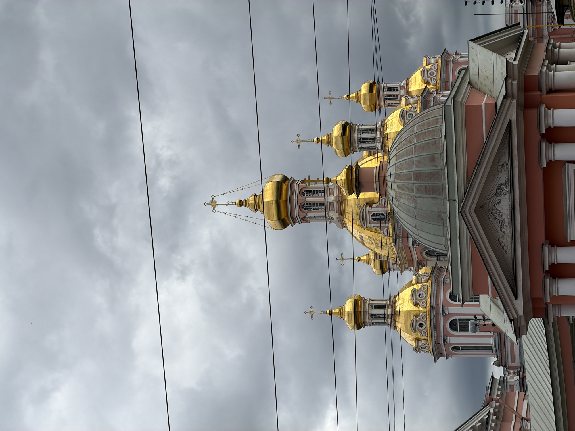 The image shows a view of the Catherine Church in Saint Petersburg, Russia, under a cloudy sky. The church is characterized by its multiple golden domes topped with crosses. The building exhibits pink facades with white architectural details, including columns and window frames. Several black electrical wires are visible crisscrossing the frame, obscuring parts of the sky and the upper sections of the church. In the foreground, a closer view of the lower part of the church reveals a large, dull-colored dome and a pediment adorned with relief sculptures. A single person in a white hard hat is visible on a balcony in the middle ground, looking towards the right.