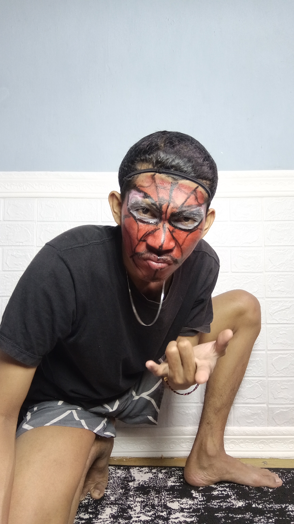 A young Indonesian male, posing in a squatting position, has applied red and black face paint in a Spider-Man-like design. He is wearing a black t-shirt and grey patterned shorts. The background consists of a white textured wall and a light blue wall above it. The image was taken indoors, likely in a residential setting in Kebumen, Indonesia.